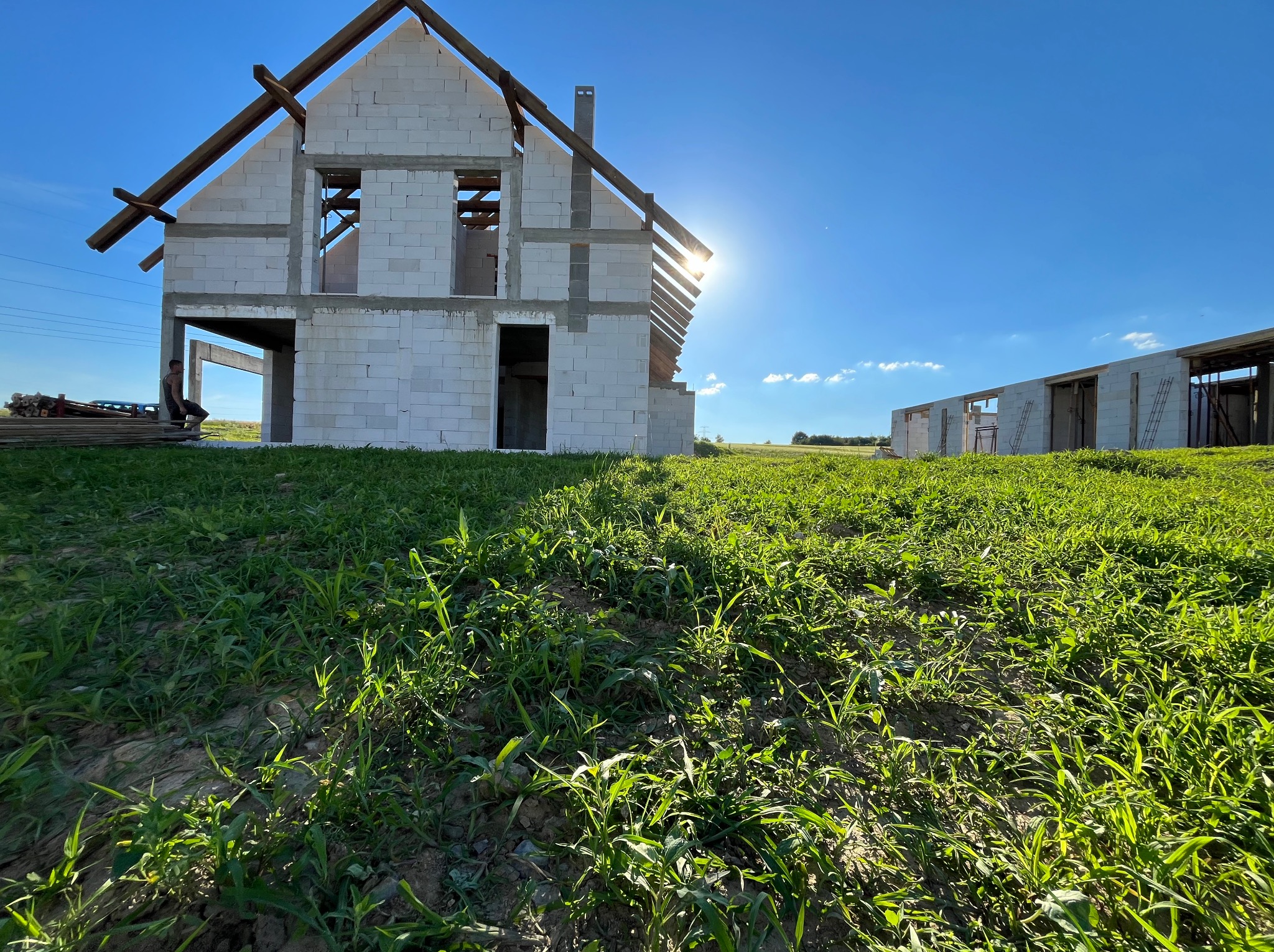 Wznoszona konstrukcja dwukondygnacyjnego budynku z białych bloczków, widoczne drewniane elementy więźby dachowej, w tle podobny budynek w trakcie budowy, zielona łąka i błękitne niebo z promieniami...