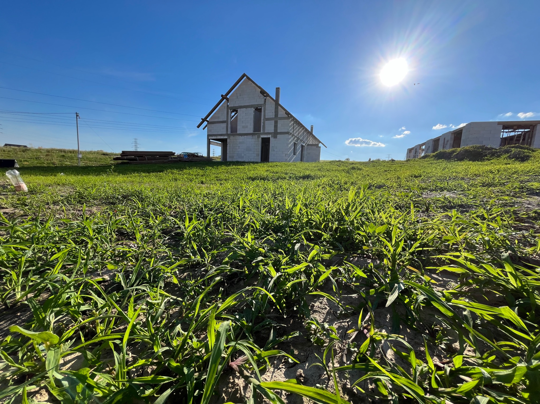Wznoszący się na tle błękitnego nieba budynek w stanie surowym otwartym, z widoczną konstrukcją dachu i ścianami z betonu komórkowego, sfotografowany z perspektywy niskiej trawy.