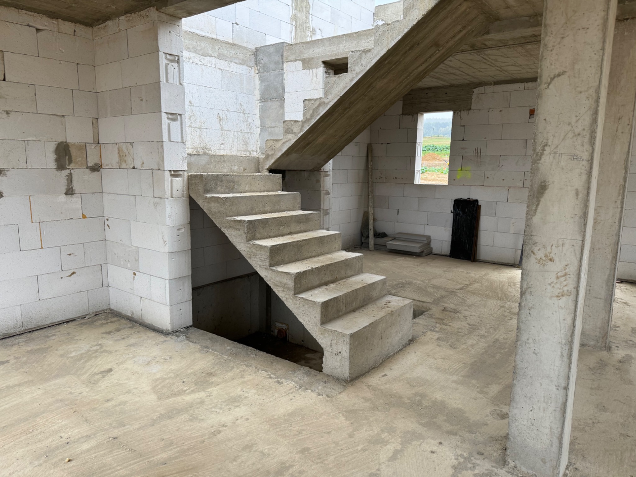 Surowe wnętrze w trakcie budowy z betonowymi schodami, mury z białych bloczków, widoczne okno z zielonym krajobrazem w tle oraz betonowe filary.