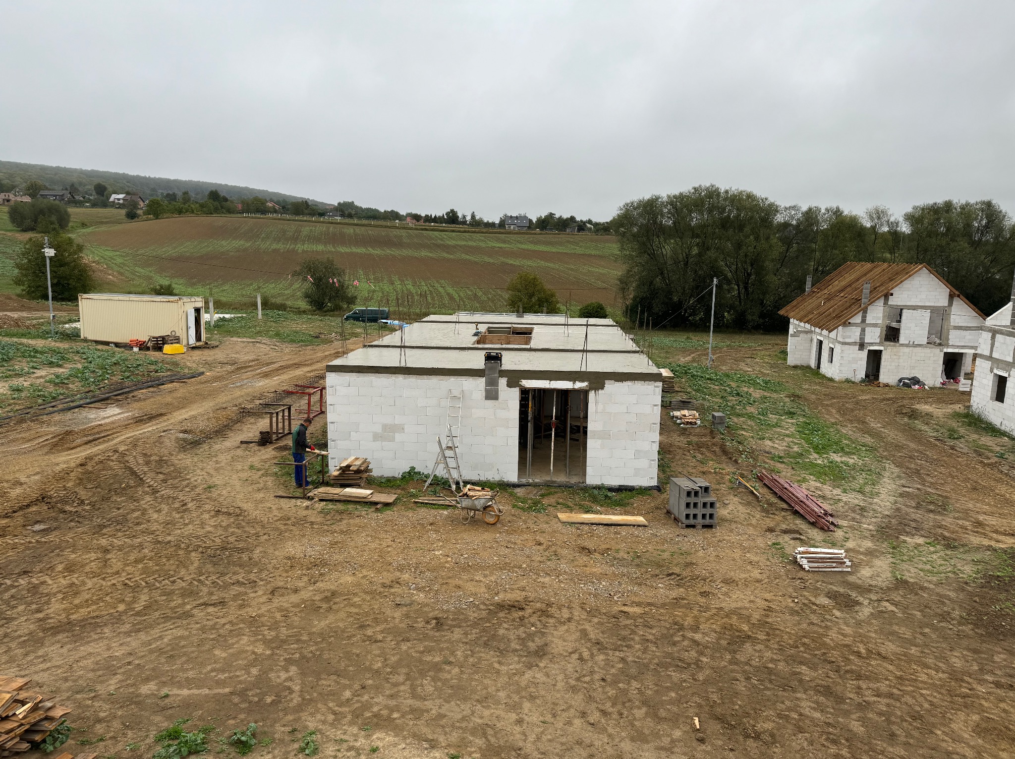 Budowa domu z betonu komórkowego w stanie surowym otwartym, widok z góry na plac budowy z pracującym robotnikiem, w tle pola i zabudowa wiejska w pochmurny dzień.