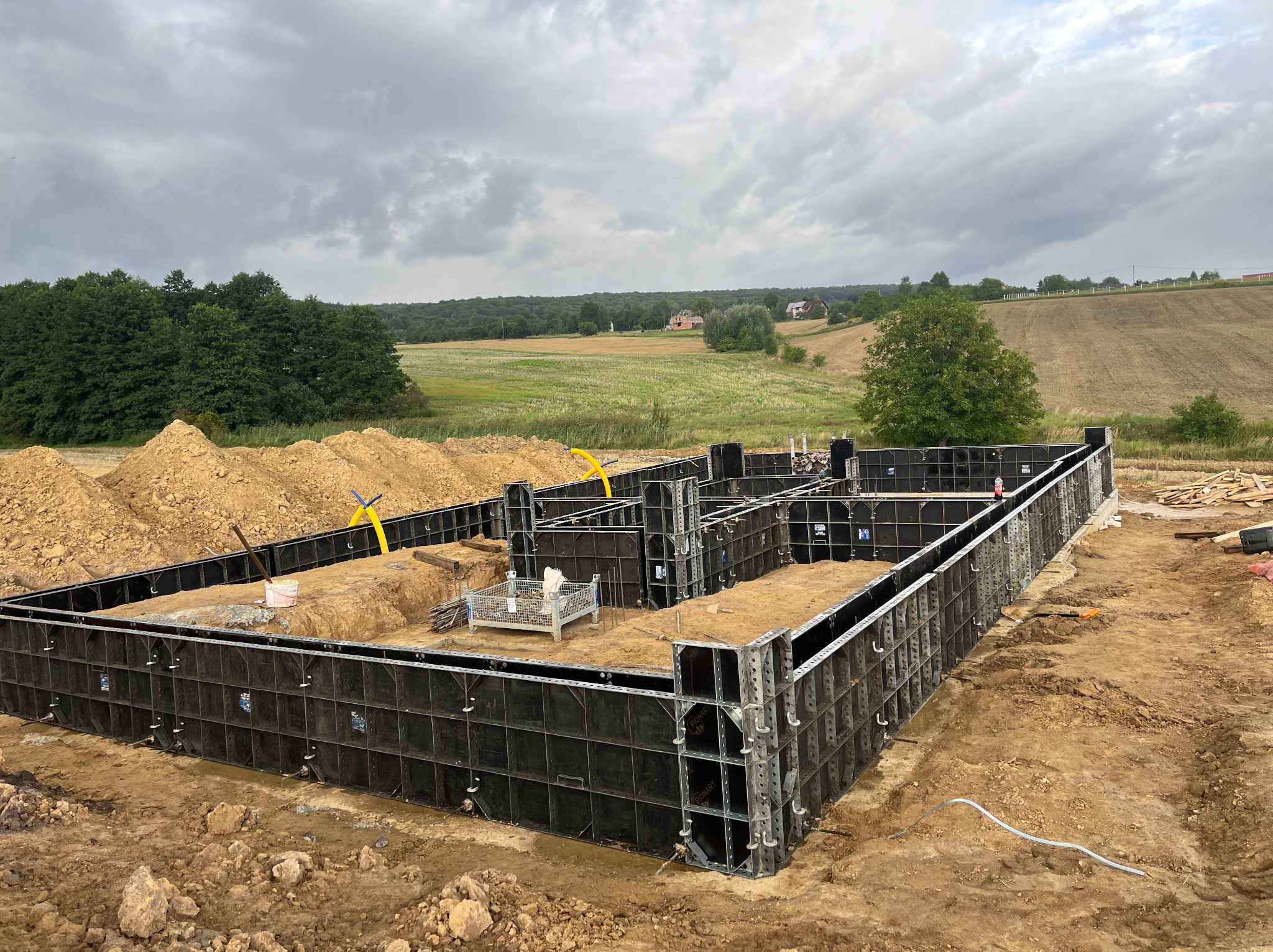 Szare, pochmurne niebo nad przygotowanym szalunkiem fundamentów domu jednorodzinnego na tle krajobrazu z polami i lasami.