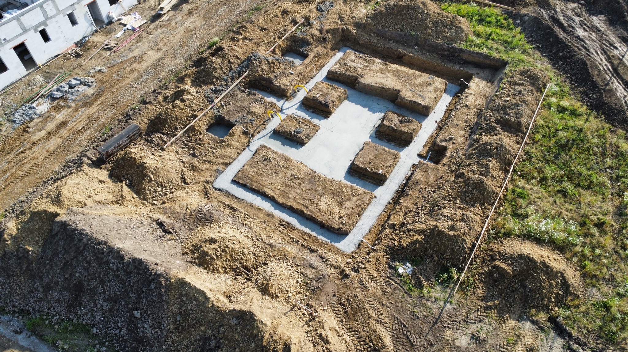 Widok z drona na wykop pod fundamenty z wylanym betonem, widoczne wystające przewody, w tle budynek w stanie surowym otwartym.
