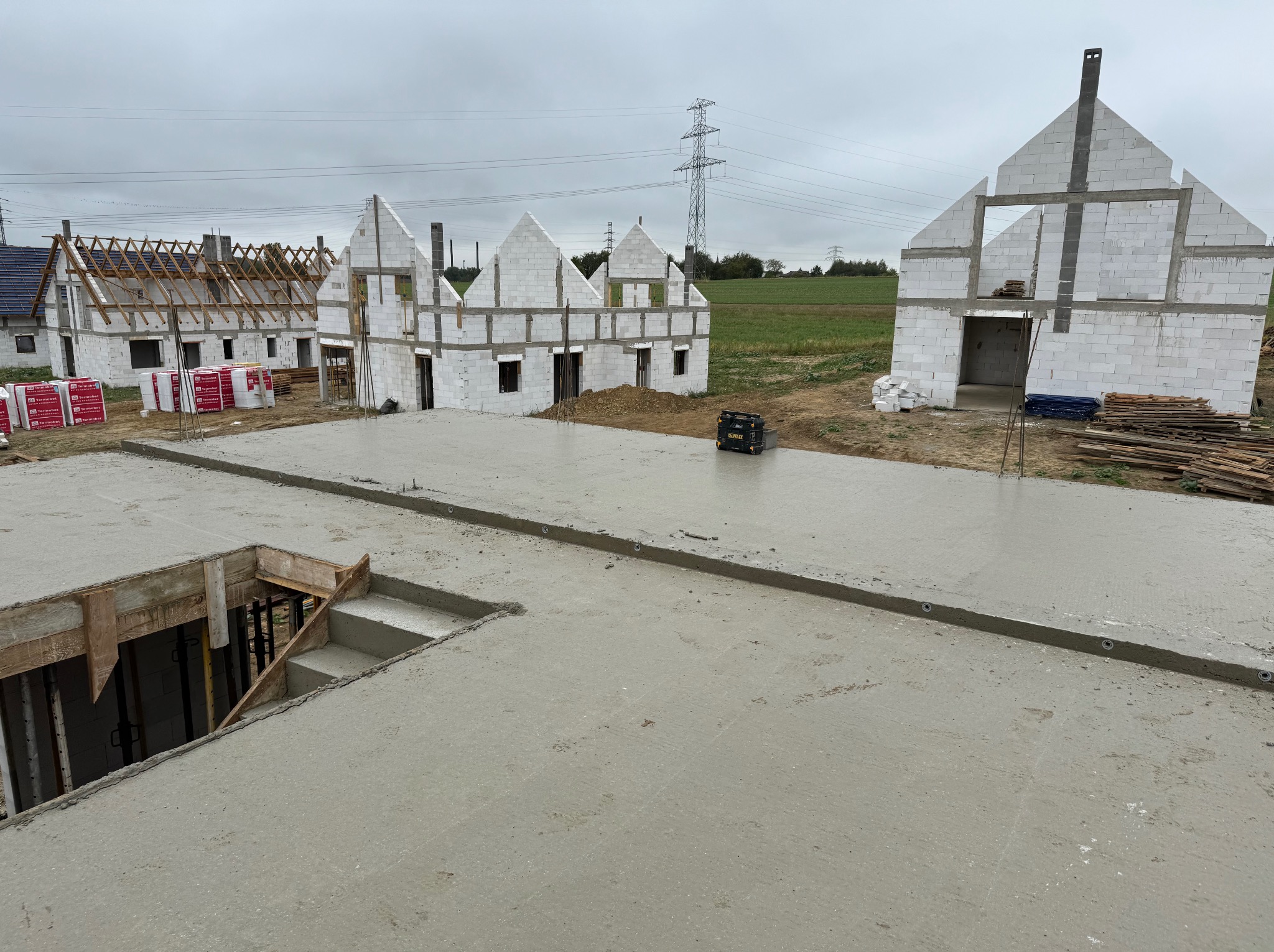 Wylana płyta fundamentowa z widocznym otworem na schody, w tle budowa kilku domów z białych bloczków i jeden z drewnianą konstrukcją dachu.