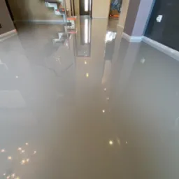 Szara, błyszcząca posadzka żywiczna w nowym domu, odbijająca schody i światło z okien, z zabezpieczonymi folią ścianami w trakcie wykończenia.