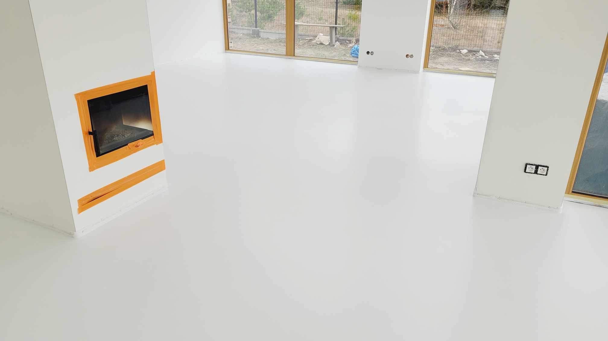 Biała, lśniąca posadzka w minimalistycznym wnętrzu z kominkiem obudowanym pomarańczową ramą i dużymi oknami z drewnianymi ramami. Widoczne gniazdka elektryczne na ścianach.