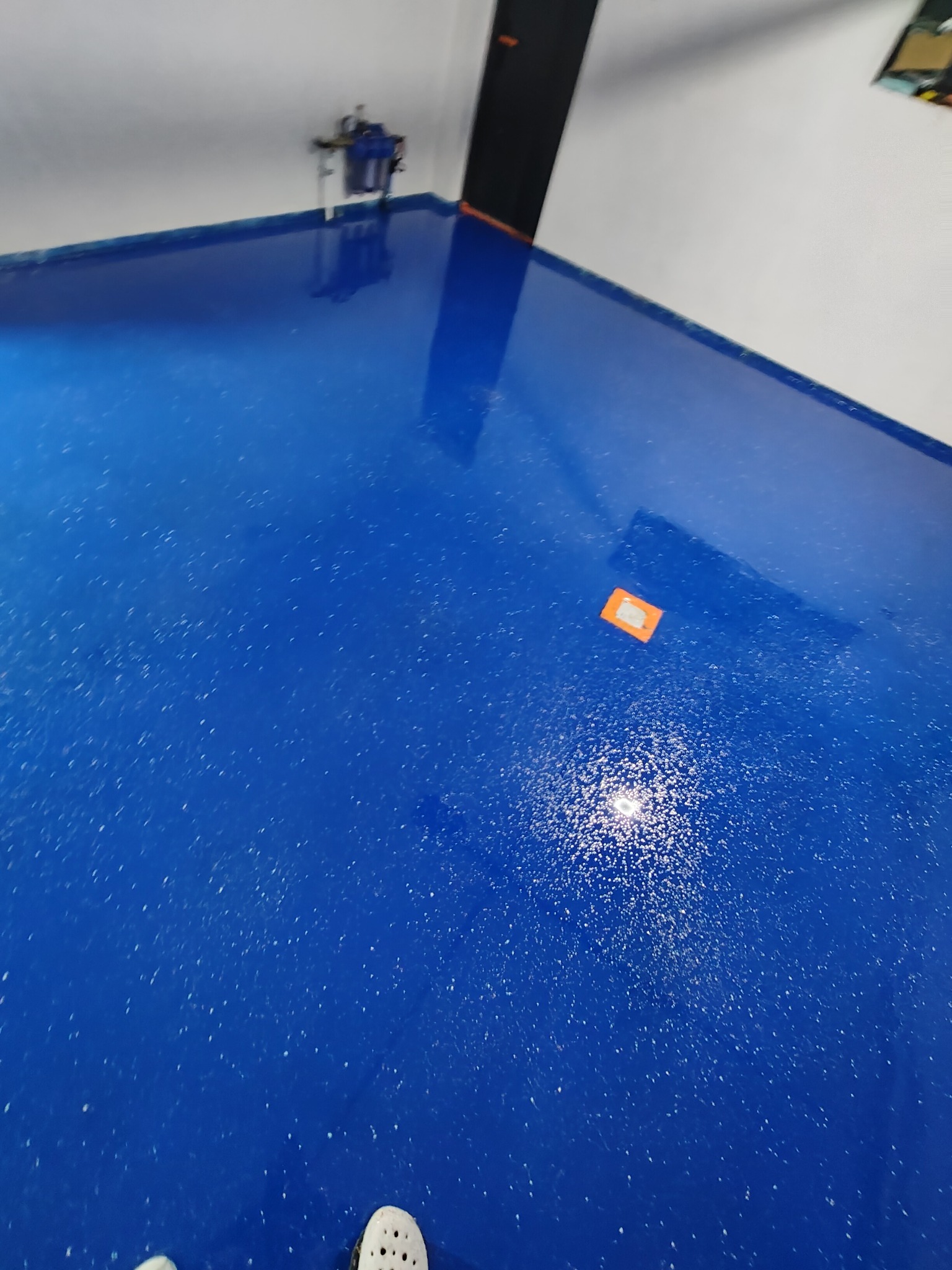 Błyszcząca, intensywnie niebieska posadzka z żywicy z widocznymi drobnymi białymi inkluzjami, odbijająca światło w centralnej części, z pomarańczową naklejką w tle i fragmentem białego buta...