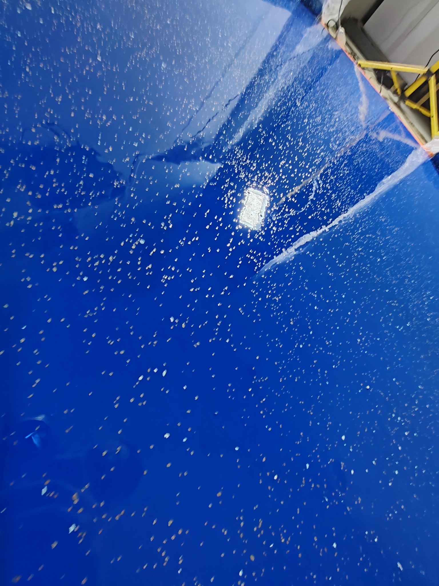 Błyszcząca, błękitna posadzka żywiczna z zatopionymi drobinkami brokatu, odbijająca światło lampy sufitowej, widoczny fragment rusztowania przy ścianie.