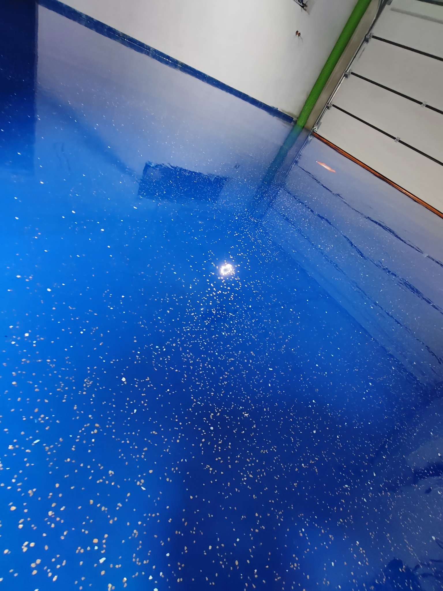 Błyszcząca, błękitna podłoga z żywicy z drobnymi, jasnymi wtrąceniami, widziana pod kątem w rogu pomieszczenia z białą ścianą i fragmentem bramy garażowej.