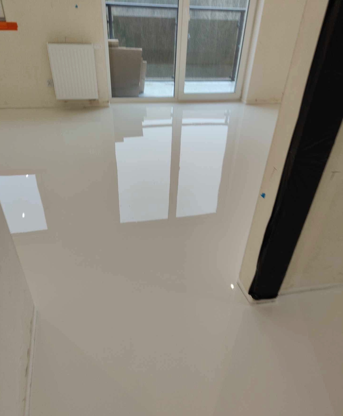 Błyszcząca, jasna podłoga w pokoju z dużym oknem balkonowym, widoczne odbicia światła i elementów otoczenia, zabezpieczona taśmą malarską przy ścianie.