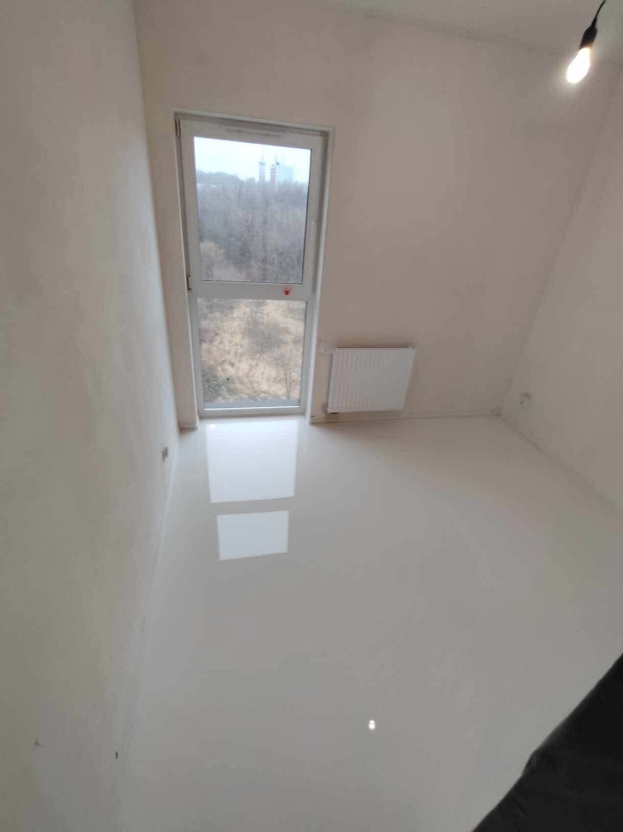 Biała, błyszcząca posadzka w pustym pokoju w Tomaszowie Mazowieckim, odbijająca światło z okna i lampy sufitowej, świeżo po remoncie.