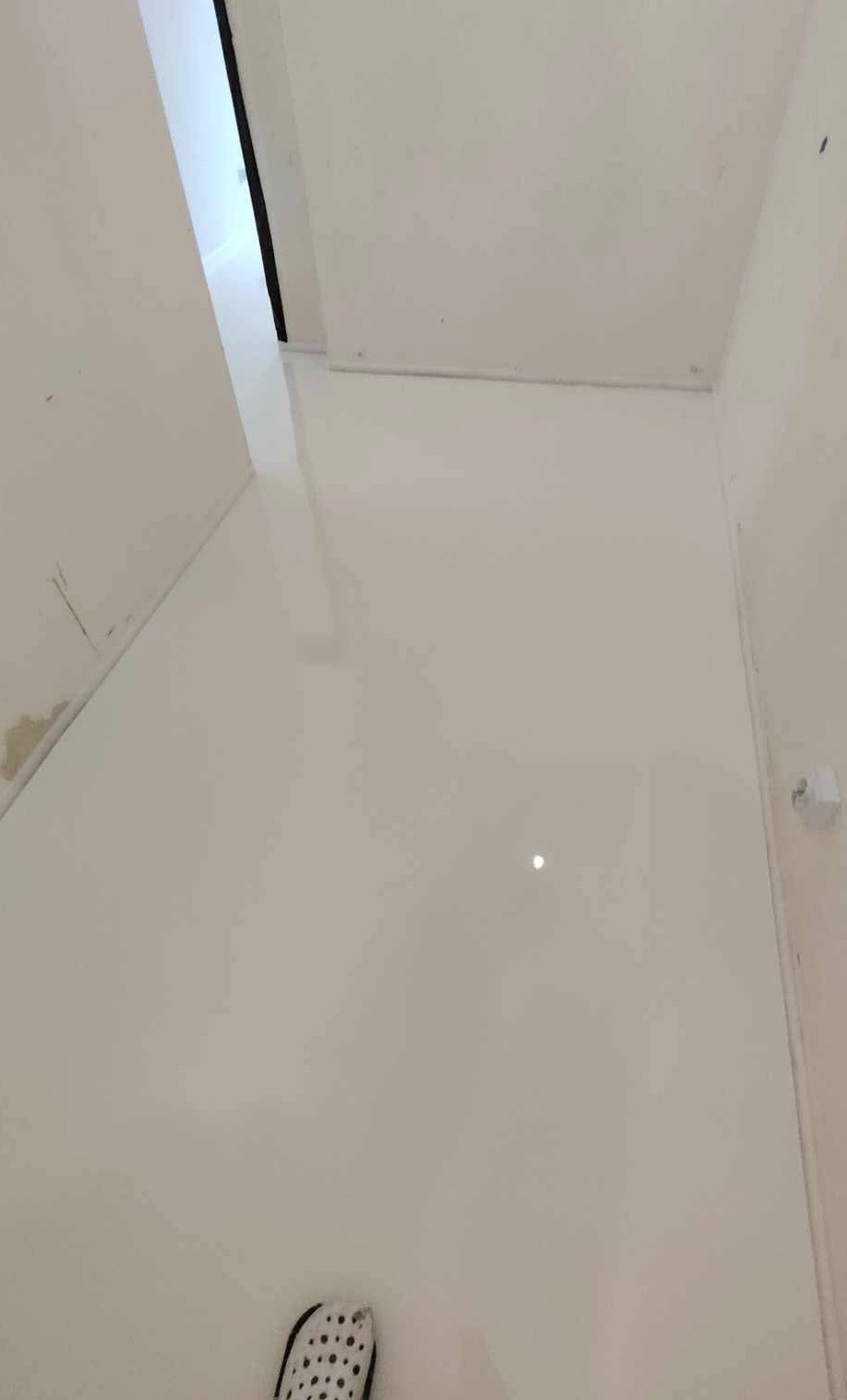 Błyszcząca, biała podłoga w rogu pomieszczenia z widocznym fragmentem buta w lewym dolnym rogu, sugerująca świeżo wylaną powierzchnię.