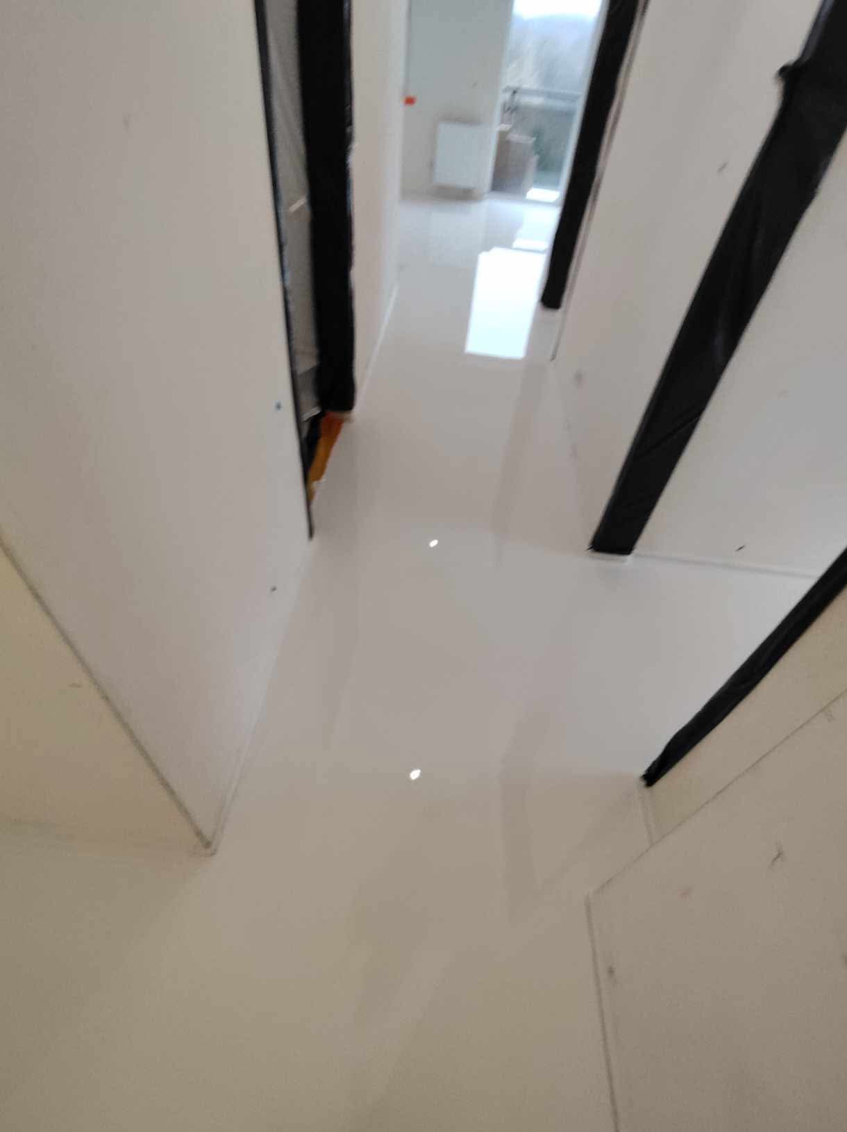 Błyszcząca, biała podłoga w korytarzu, zabezpieczona czarną folią przy ścianach, widok w głąb z oknem na końcu.