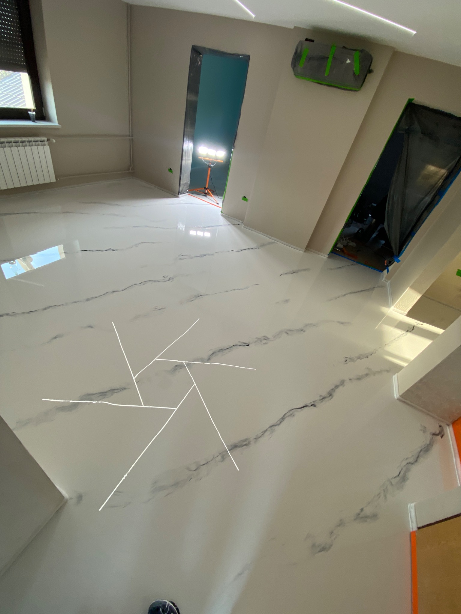 Błyszcząca, biała podłoga z żywicy epoksydowej imitująca marmur, z widocznymi ciemnymi żyłkami, w trakcie prac wykończeniowych; zabezpieczone folią framugi drzwi, oświetlenie budowlane w tle.