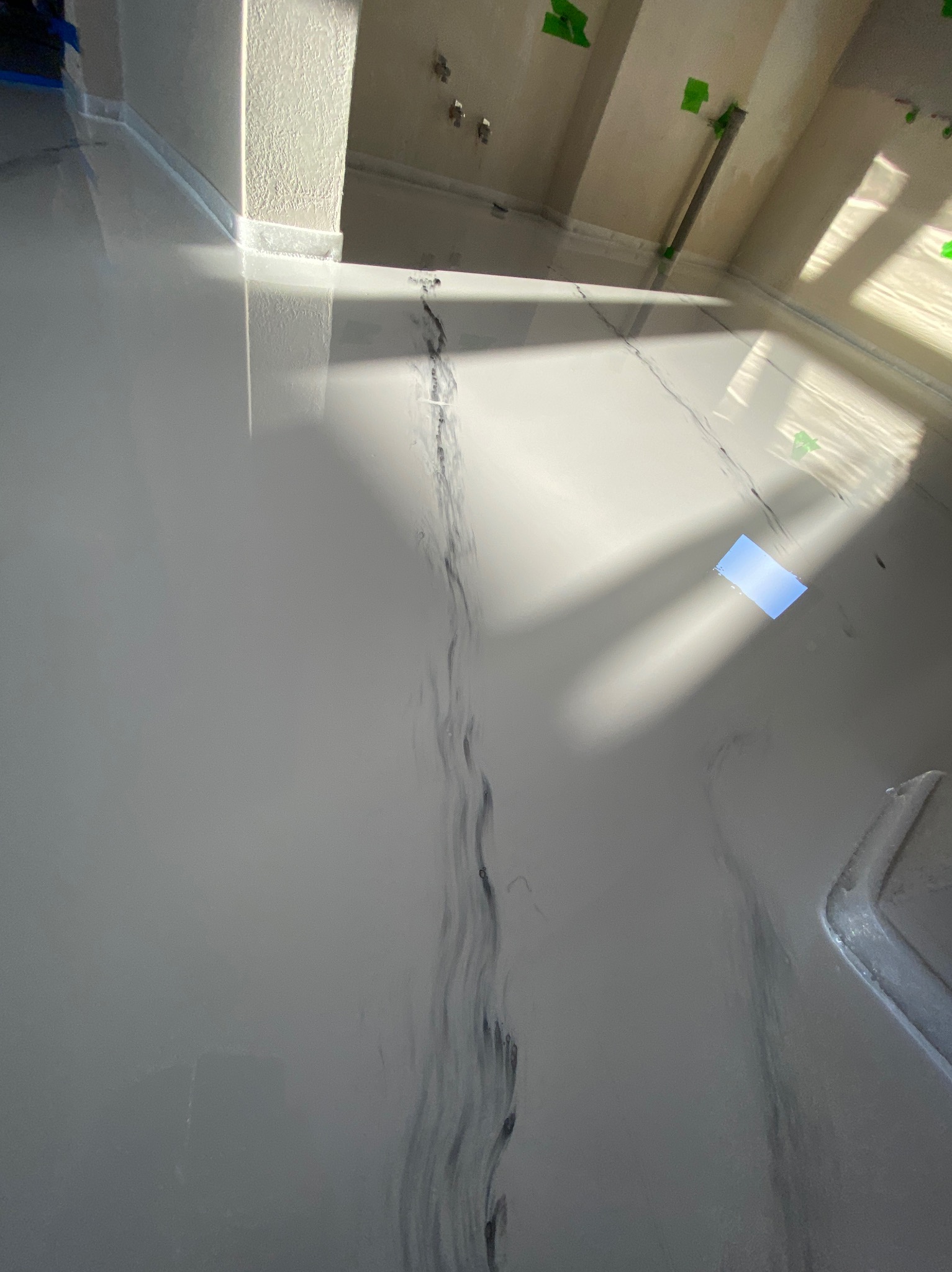 Świeżo wylana, jasnoszara posadzka z żywicy z ciemnymi smugami, mokra i odbijająca światło, widoczne zielone taśmy malarskie na ścianach i rurach, w tle wpadające promienie słoneczne.