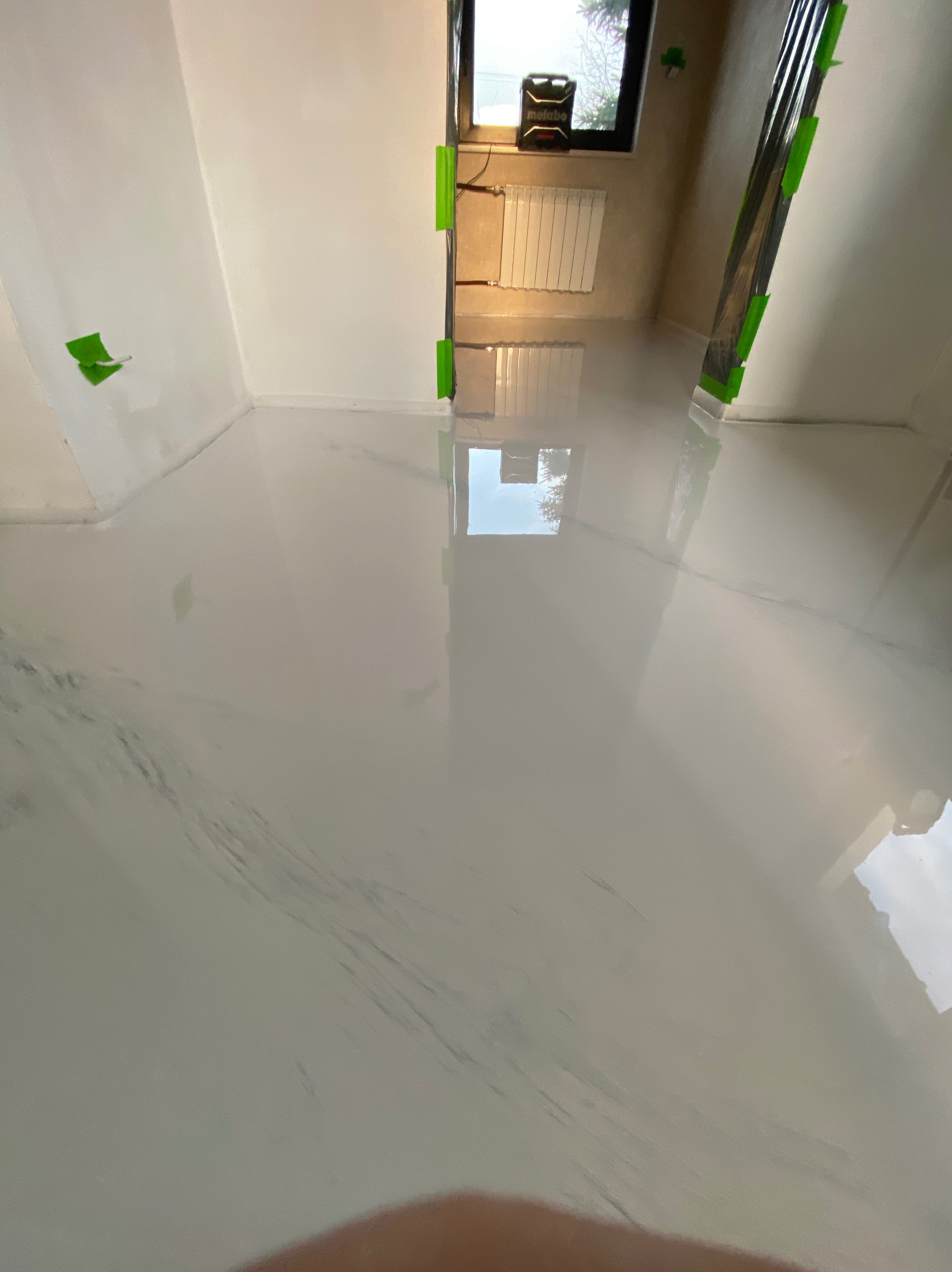 Biała, błyszcząca podłoga z żywicy epoksydowej w trakcie schnięcia, zabezpieczone folią ściany z zieloną taśmą malarską, widoczny grzejnik i okno z odbijającym się niebem.