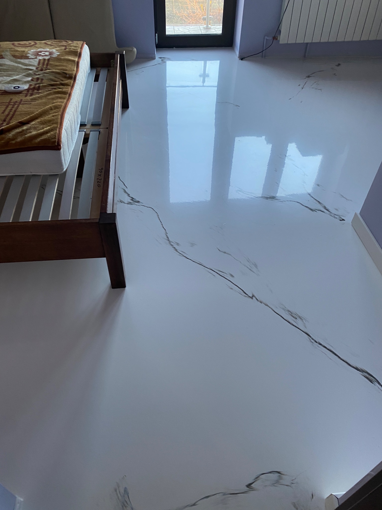 Błyszcząca, biała podłoga z żywicy epoksydowej imitująca marmur w sypialni, odbijająca światło okna i fragment łóżka z materacem.