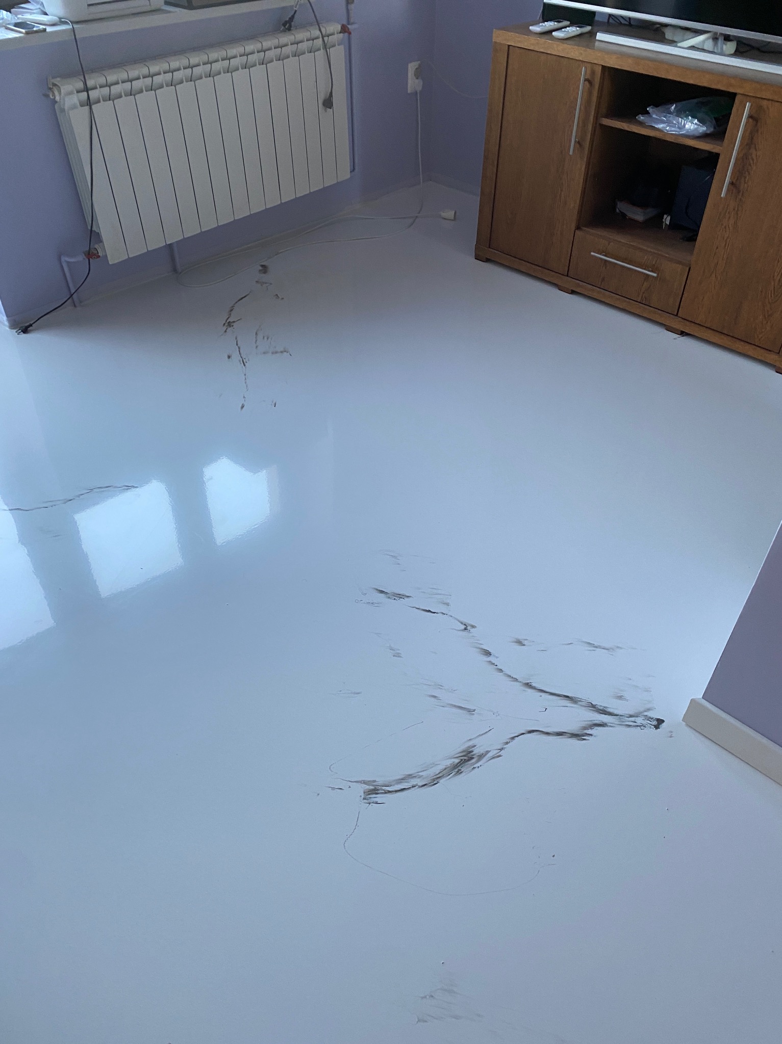 Błyszcząca, jasna podłoga z widocznymi smugami zabrudzeń i śladami niedoczyszczeń, odbijająca światło okna, w tle grzejnik i drewniana komoda.