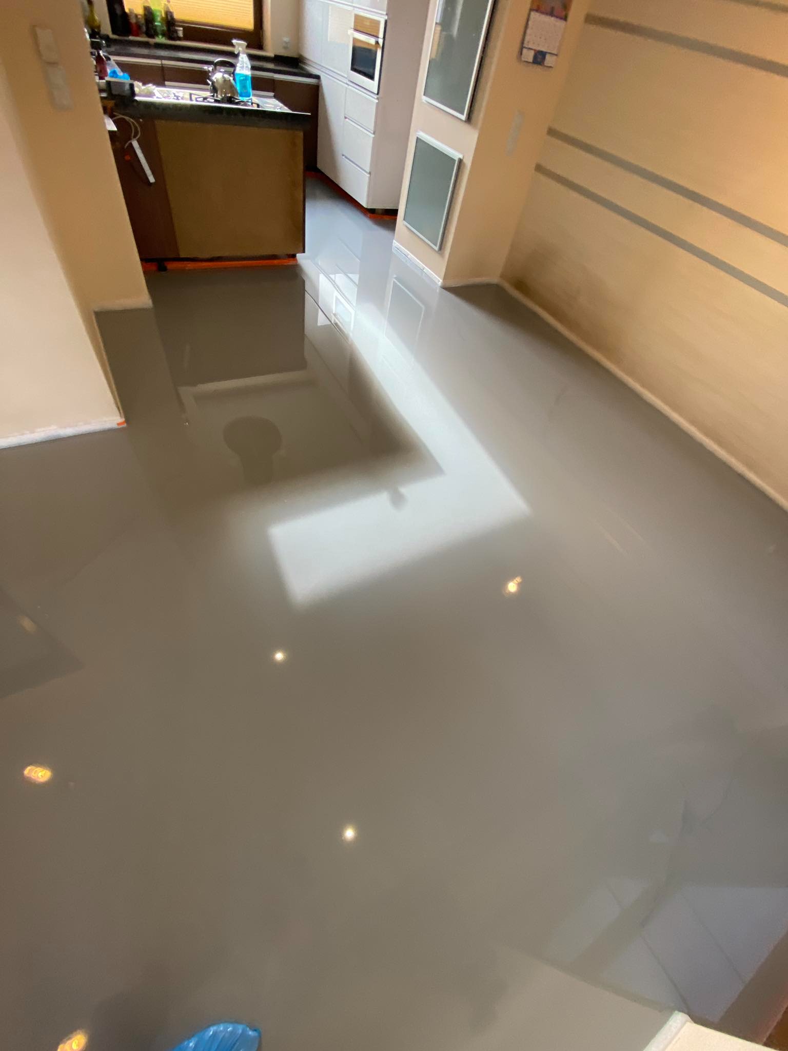 Błyszcząca, szara posadzka żywiczna w nowocześnie wykończonym wnętrzu, z widocznymi refleksami światła i fragmentem zabudowy kuchennej w tle.