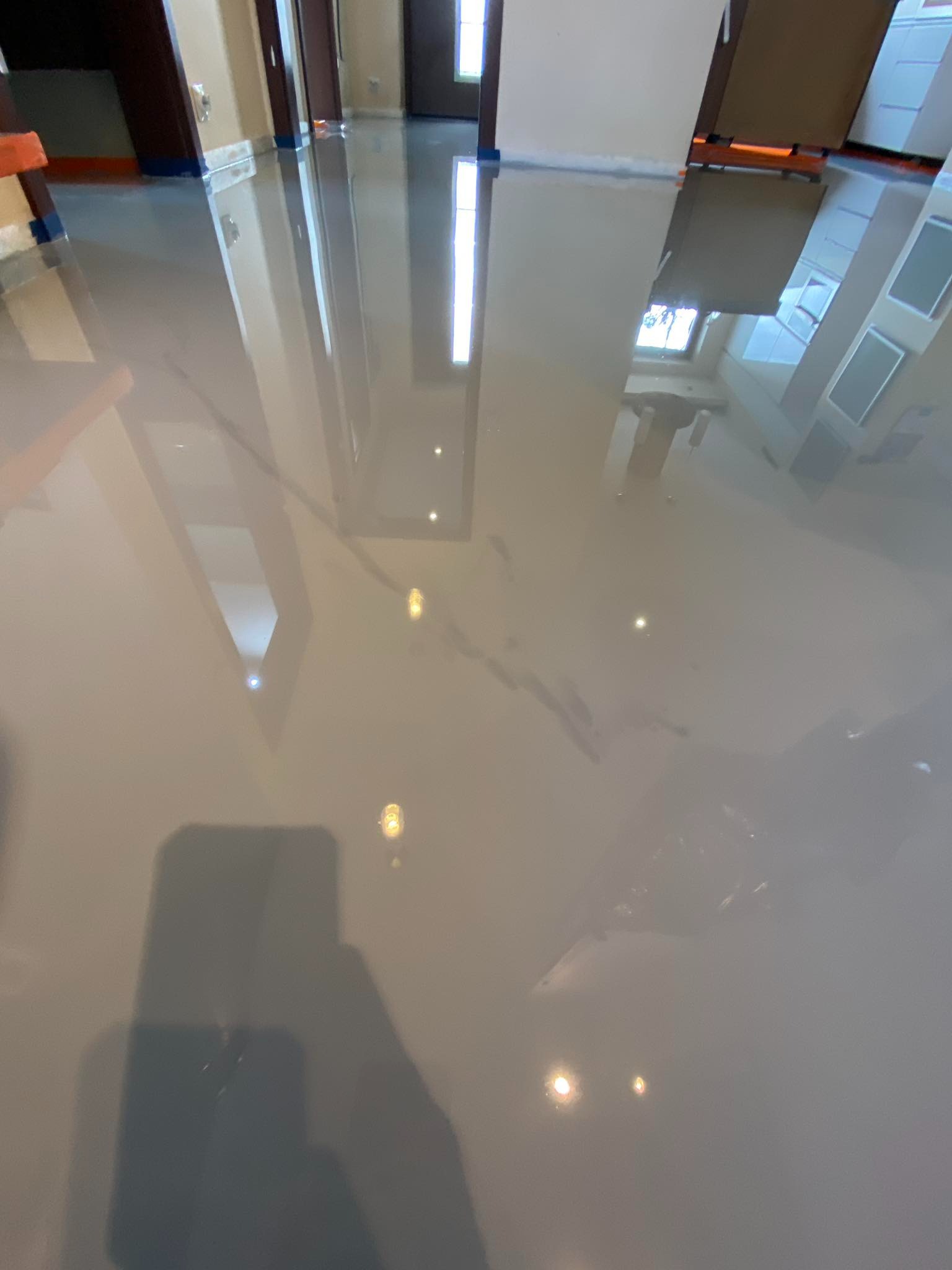 Jasnoszara, błyszcząca podłoga z żywicy epoksydowej w nowo wykończonym wnętrzu, odbijająca światło i elementy otoczenia, z widocznym cieniem osoby robiącej zdjęcie.