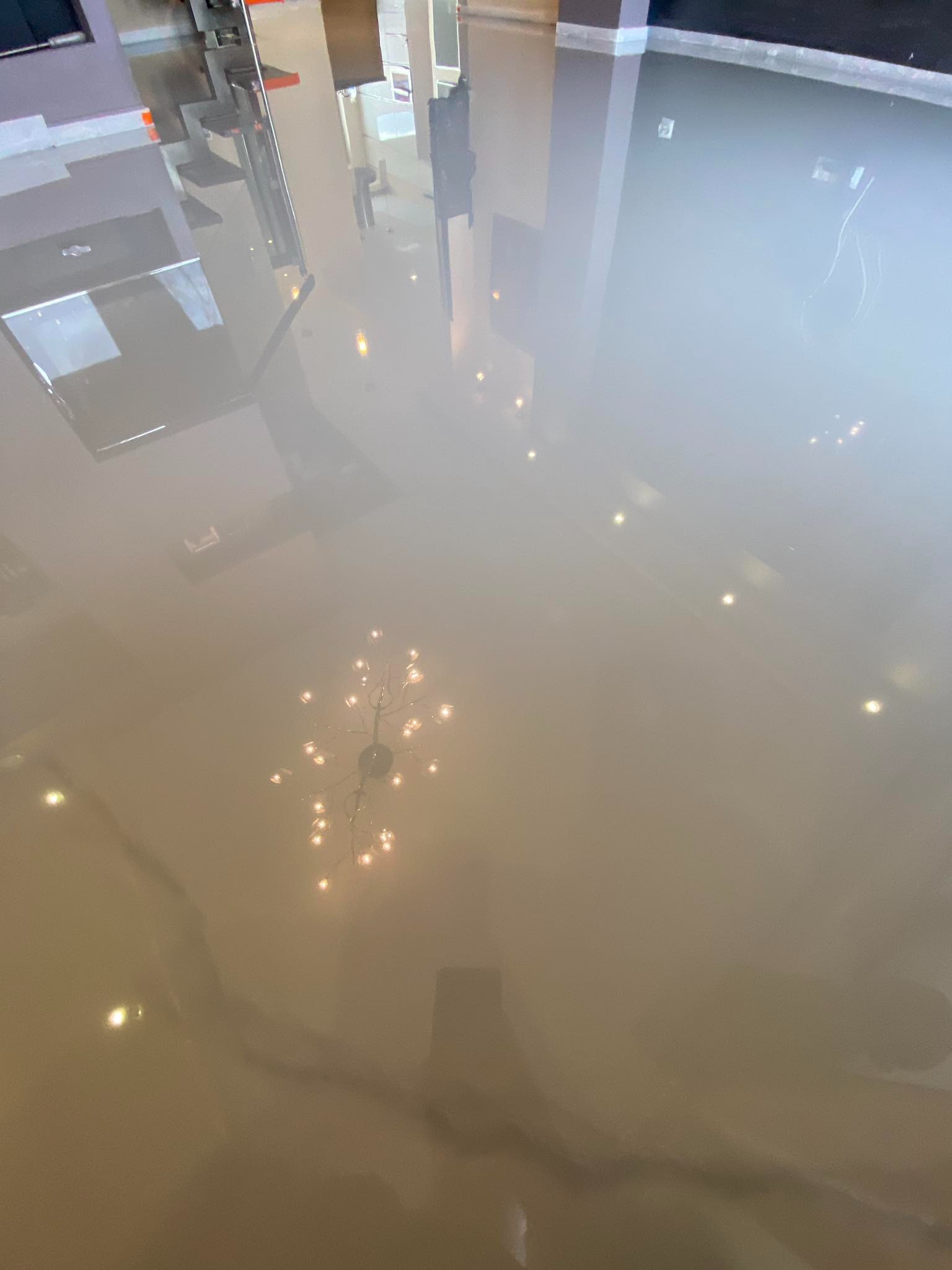 Beżowa, lśniąca posadzka z żywicy epoksydowej odbijająca światło lampy sufitowej i elementy wyposażenia wnętrza. Gładka powierzchnia z efektem lustra.