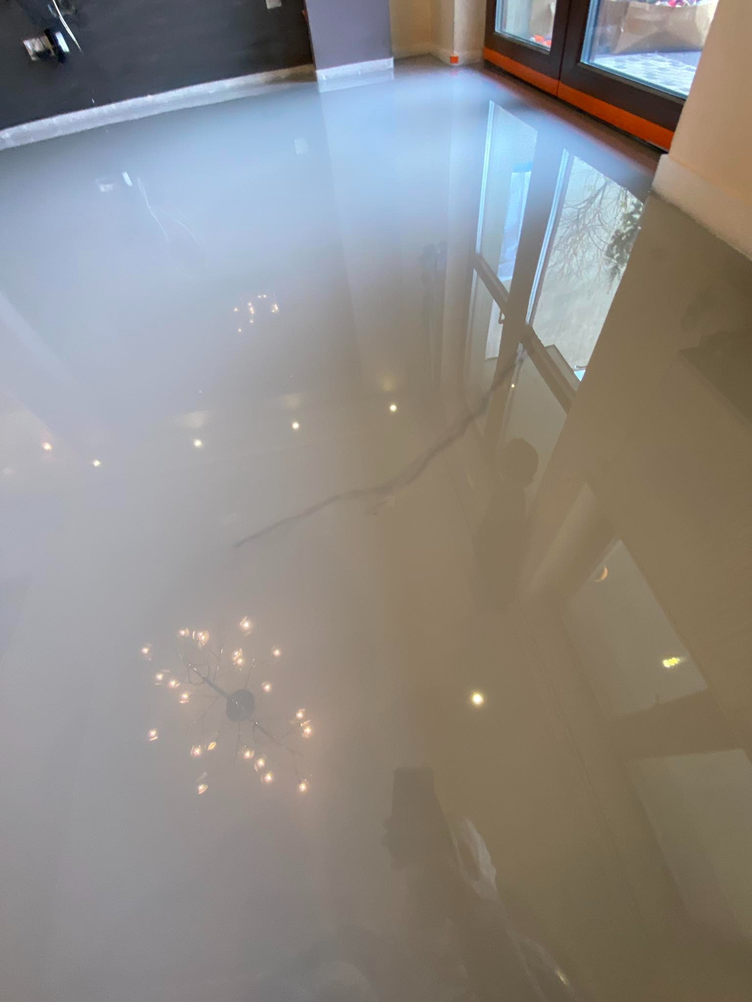 Błyszcząca, beżowa podłoga z żywicy epoksydowej, odbijająca światło żyrandola i okna balkonowego, z widoczną delikatną smugą na powierzchni.