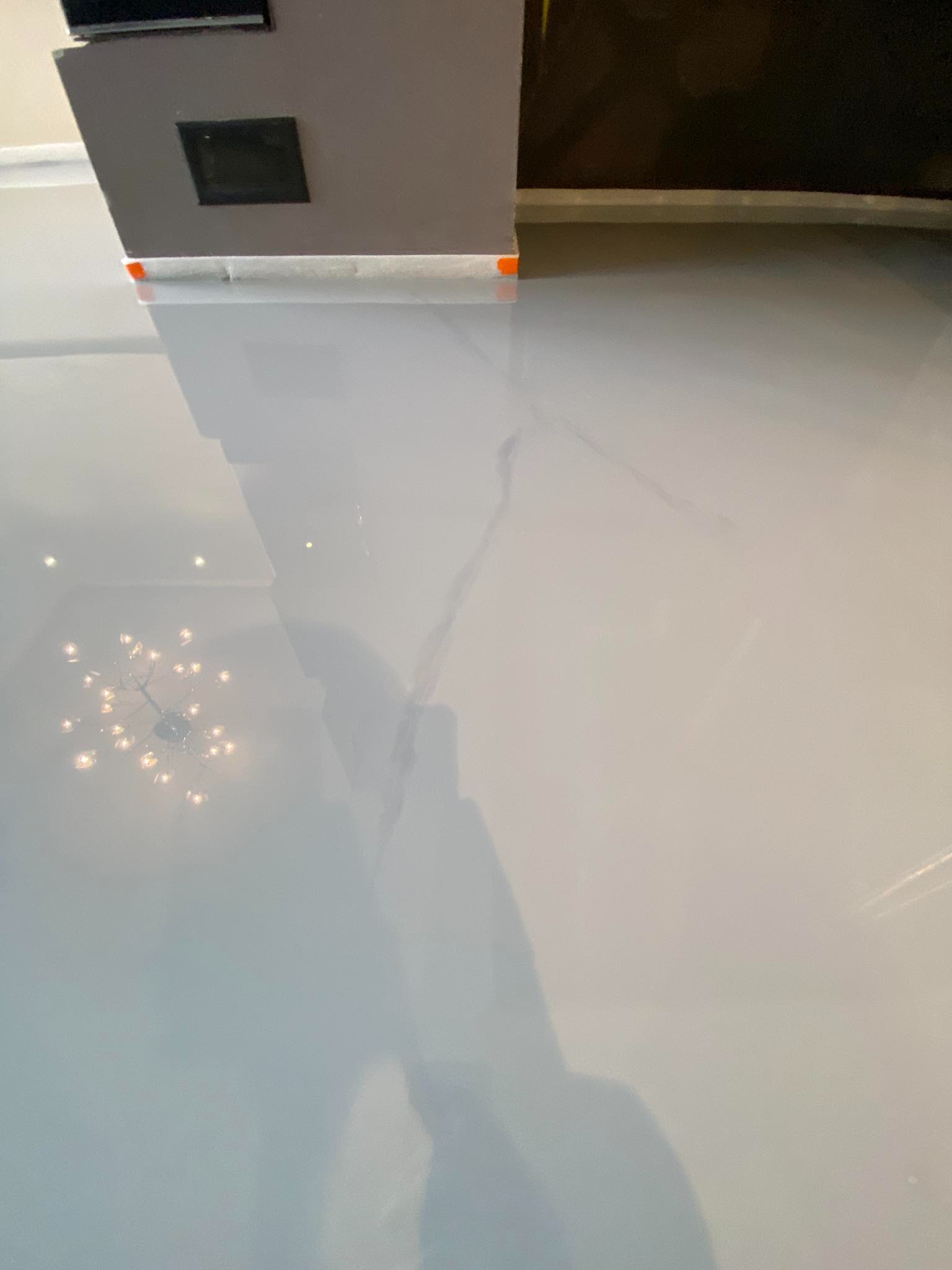 Świeżo wylana, błyszcząca, jasnoszara posadzka z żywicy epoksydowej odbija światło żyrandola i fragment mebla z czarnym elementem wentylacyjnym.