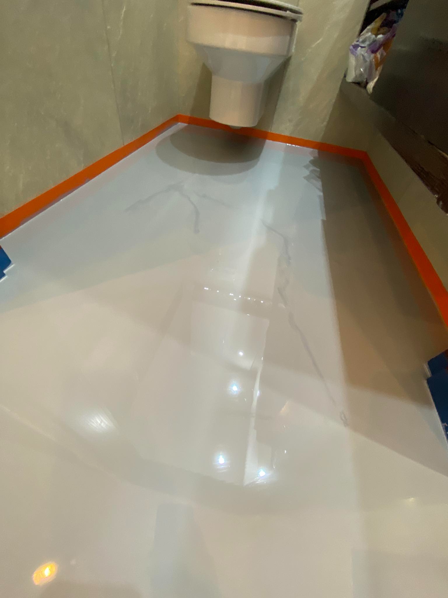 Biała, błyszcząca posadzka w łazience w trakcie wylewania, zabezpieczona pomarańczową taśmą, widoczny fragment toalety i ściany z jasnego kamienia.
