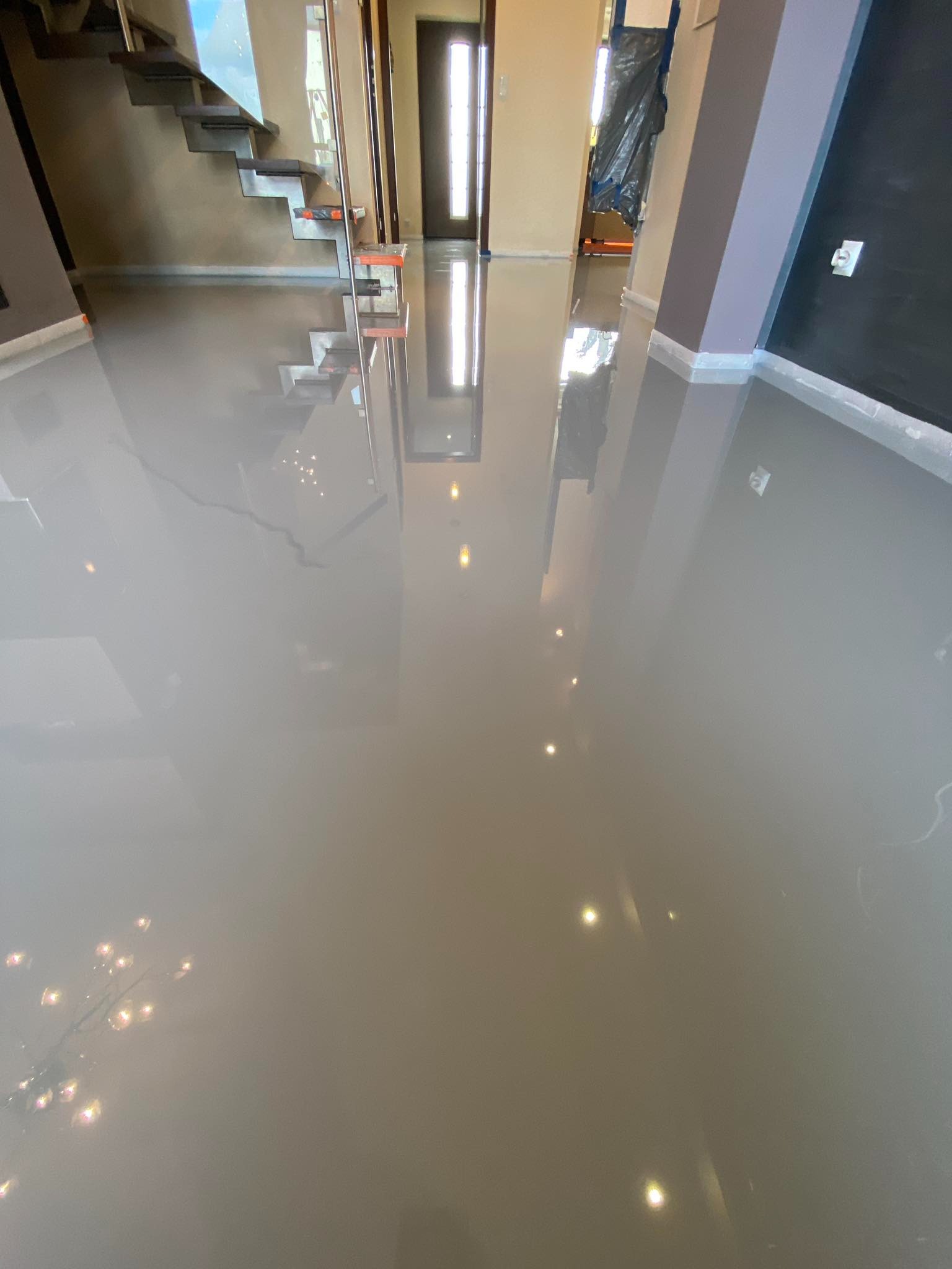 Szara, błyszcząca posadzka żywiczna w nowym domu, odbijająca schody i światło z okien, z zabezpieczonymi folią ścianami w trakcie wykończenia.