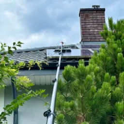 Szczotka na teleskopowym kiju czyści panele słoneczne na dachu domu z kominem, widok z dołu na tle pochmurnego nieba i zieleni drzew