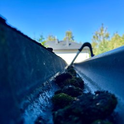 Firma Handlowo Usługowa ŁUKASZ SZLASKI - Zanieczyszczona rynna dachowa wypełniona mchem i liśćmi, narzędzie do czyszczenia rynien w tle, widok z wnętrza rynny, błękitne niebo w tle.