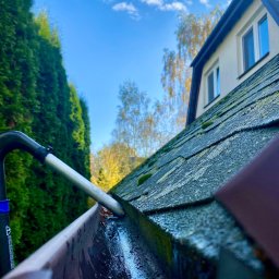 Firma Handlowo Usługowa ŁUKASZ SZLASKI - Czyszczenie rynny z poziomu dachu przy użyciu narzędzia teleskopowego; widoczna woda i zanieczyszczenia w rynnie, dach pokryty mchem, w tle iglaki i błękitne niebo z chmurami.