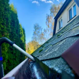 Czyszczenie rynny z poziomu dachu przy użyciu narzędzia teleskopowego; widoczna woda i zanieczyszczenia w rynnie, dach pokryty mchem, w tle iglaki i błękitne niebo z chmurami.