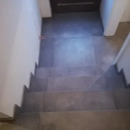 Wykończone schody wewnątrz budynku, pokryte szarą, matową glazurą, z widocznymi fragmentami instalacji wodnej przy ścianie i drzwiami w tle.