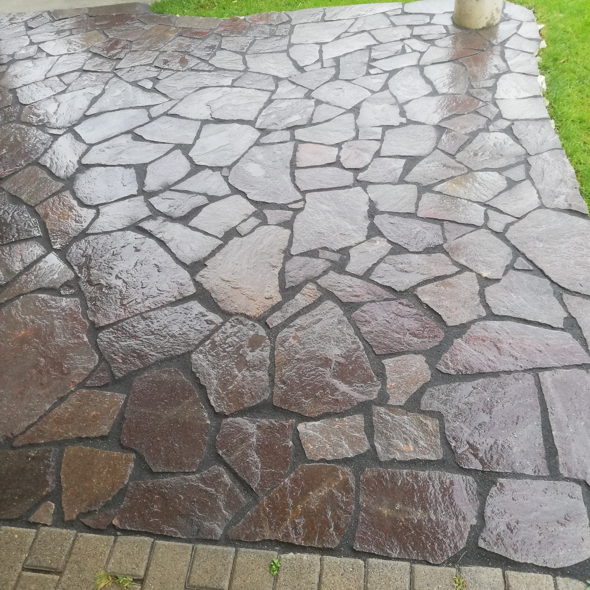 Świeżo ułożona, nieregularna nawierzchnia z kamienia naturalnego w odcieniach brązu i szarości, mokra od deszczu, z widocznymi fugami i fragmentem trawnika w tle.