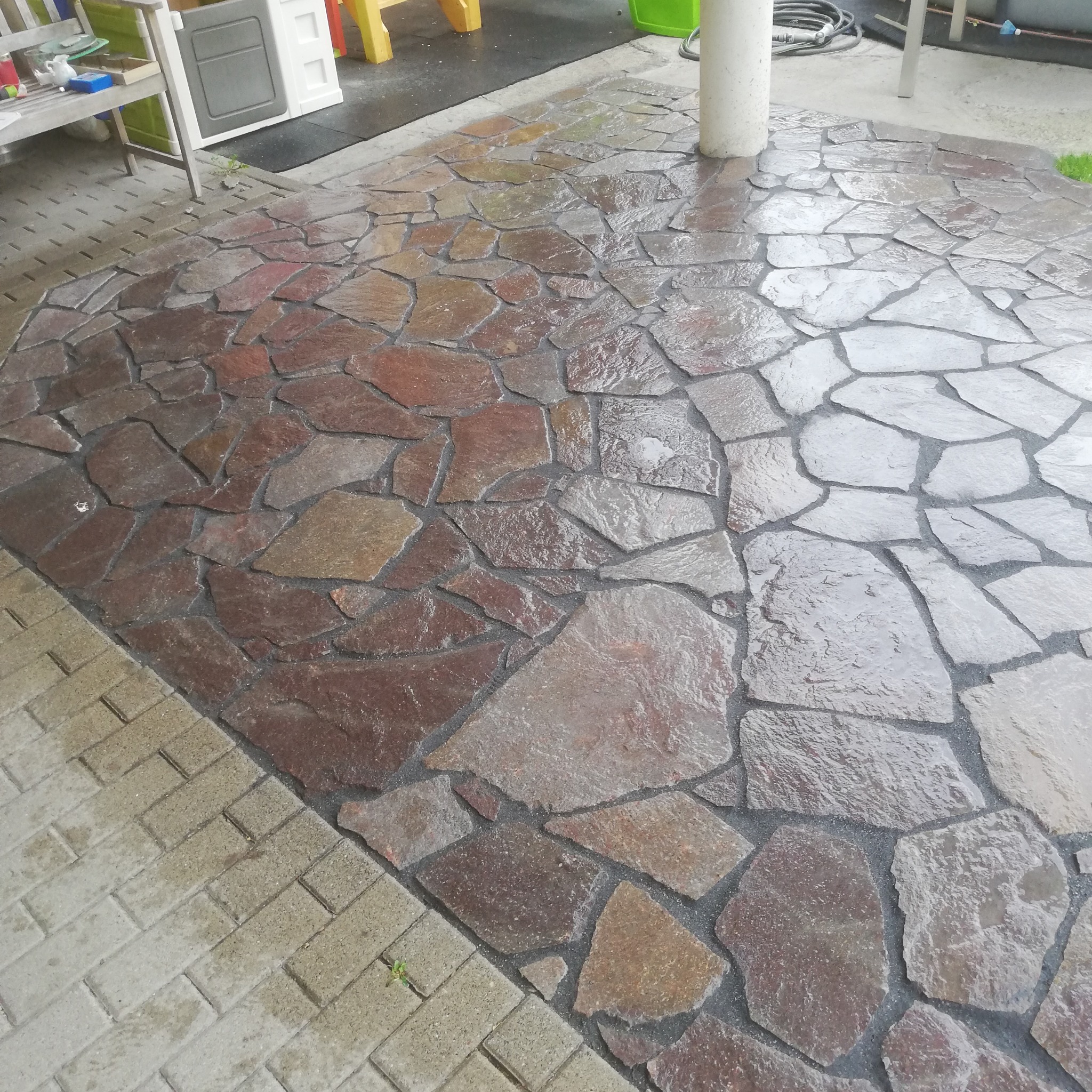 Wykończona, mokra posadzka z nieregularnych kamieni w odcieniach brązu i szarości, ułożona na zewnątrz budynku, sąsiadująca z jasną kostką brukową.