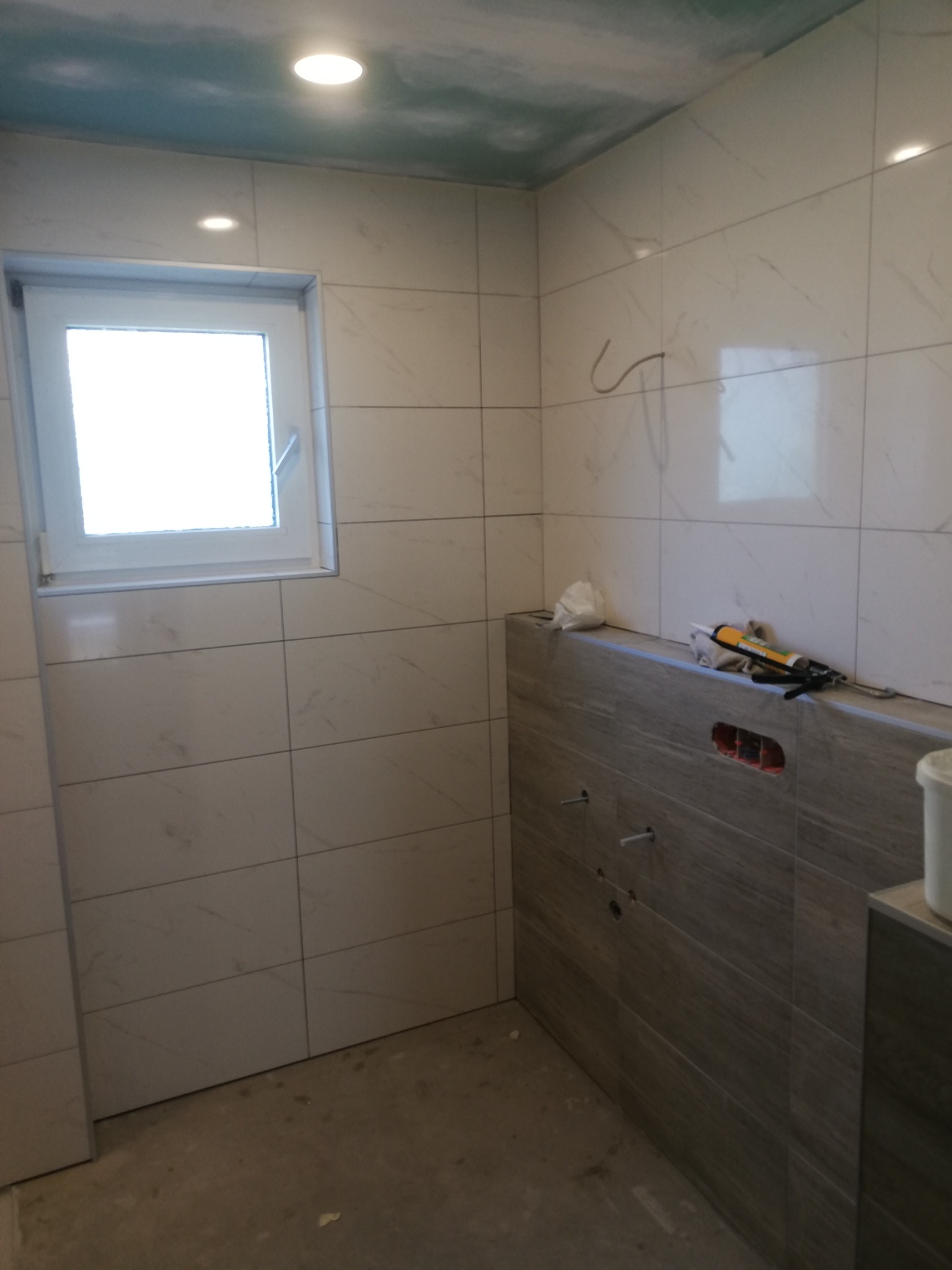 Ściany łazienki wyłożone płytkami imitującymi marmur, widoczne przygotowane przyłącza wodne i narzędzia glazurnicze na obudowie stelaża podtynkowego, sufit z efektem nieba w tle.