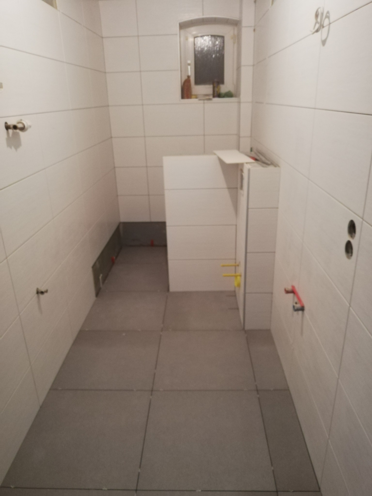 Wnętrze łazienki w trakcie prac wykończeniowych, widoczne białe płytki na ścianach, szare płytki podłogowe, fragment zabudowy stelaża WC z wystającymi elementami instalacji, małe okno w głębi...