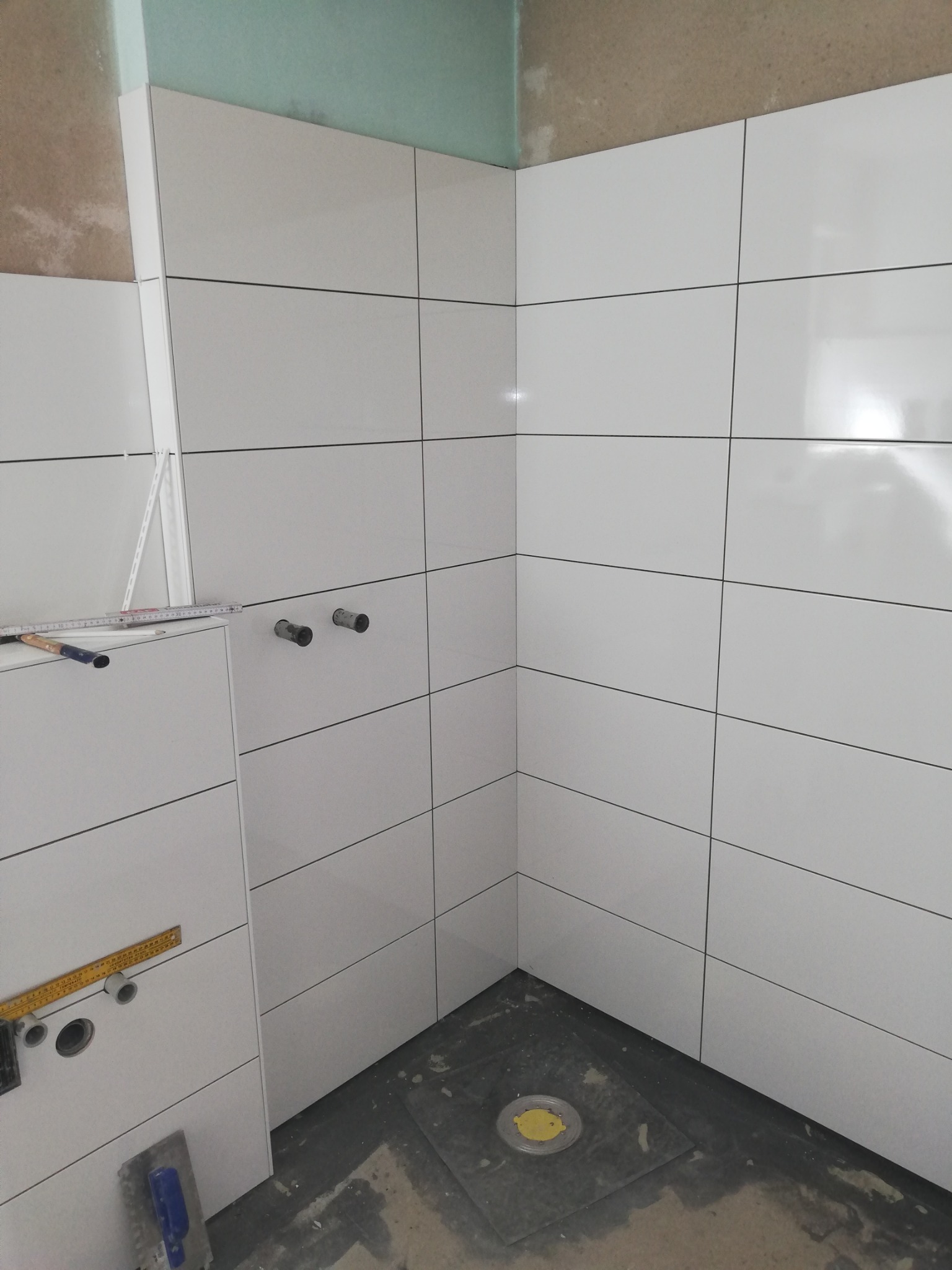 Świeżo ułożone, białe, prostokątne płytki na ścianach i zabudowie w narożniku łazienki w trakcie remontu. Widoczne wystające rury instalacji wodnej, poziomica oparta o ścianę oraz narzędzia...