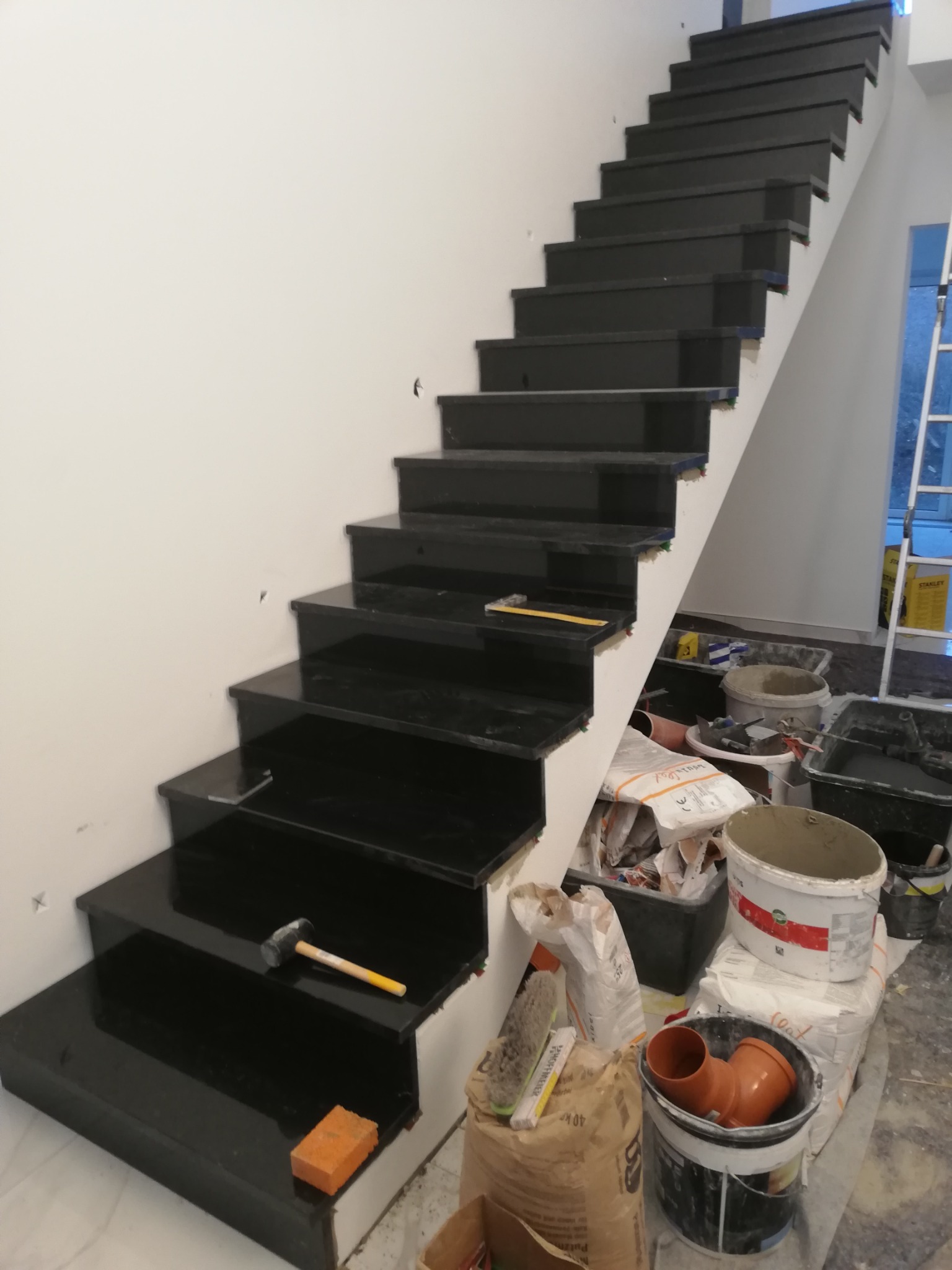 Montaż czarnych, błyszczących schodów z kamienia lub płytek, widoczne narzędzia: młotek, miarka, gąbka, kleje i wiadra z zaprawą, drabina w tle.