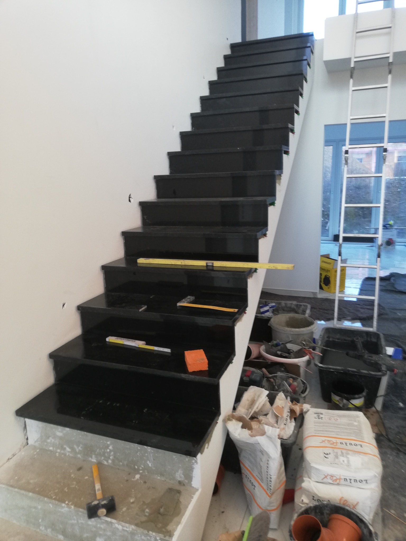 Montaż czarnych, błyszczących płytek na schodach wewnętrznych; widoczne narzędzia glazurnicze, poziomica, młotek i worki z zaprawą klejową. Schody w trakcie wykańczania, z surowymi betonowymi...