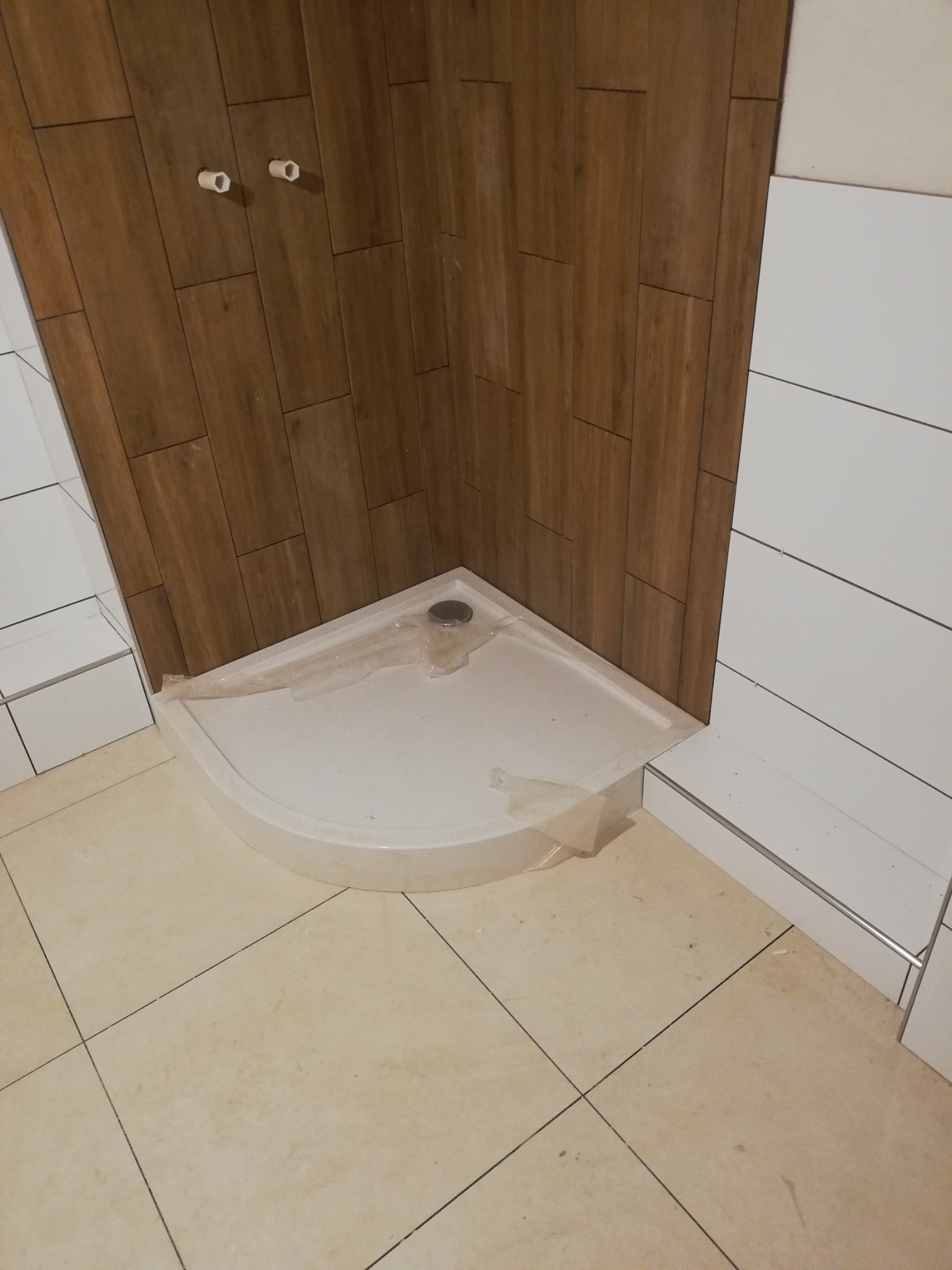 Narożna kabina prysznicowa, częściowo owinięta folią ochronną, w trakcie montażu w łazience wyłożonej płytkami imitującymi drewno i białymi, poziomymi płytkami.