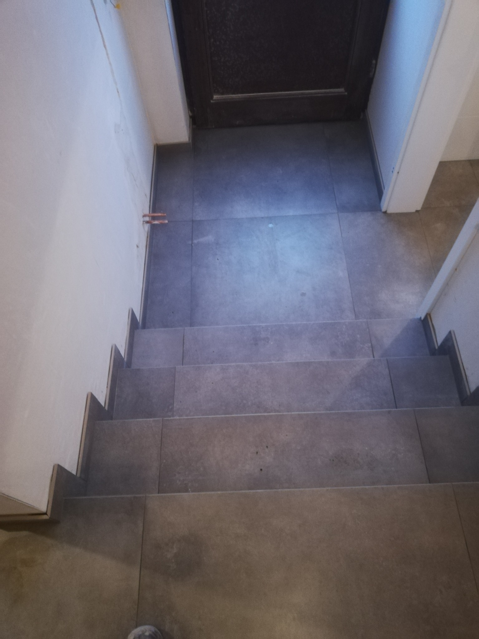 Wykończone schody wewnątrz budynku, pokryte szarą, matową glazurą, z widocznymi fragmentami instalacji wodnej przy ścianie i drzwiami w tle.