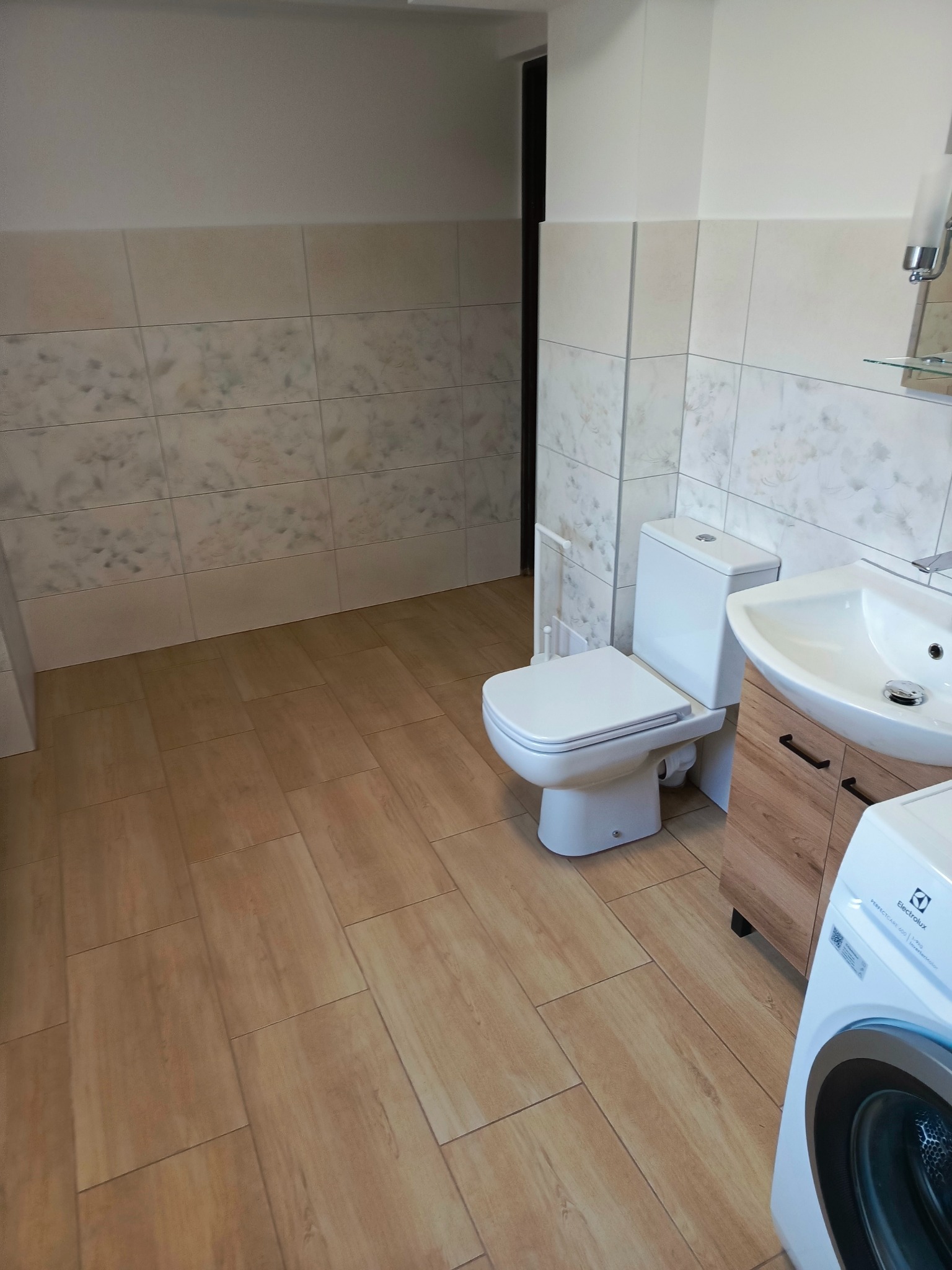 Jasna łazienka po remoncie, z beżowymi płytkami imitującymi drewno na podłodze, białą armaturą (toaleta, umywalka z szafką w kolorze drewna) i pralką Electrolux w rogu.