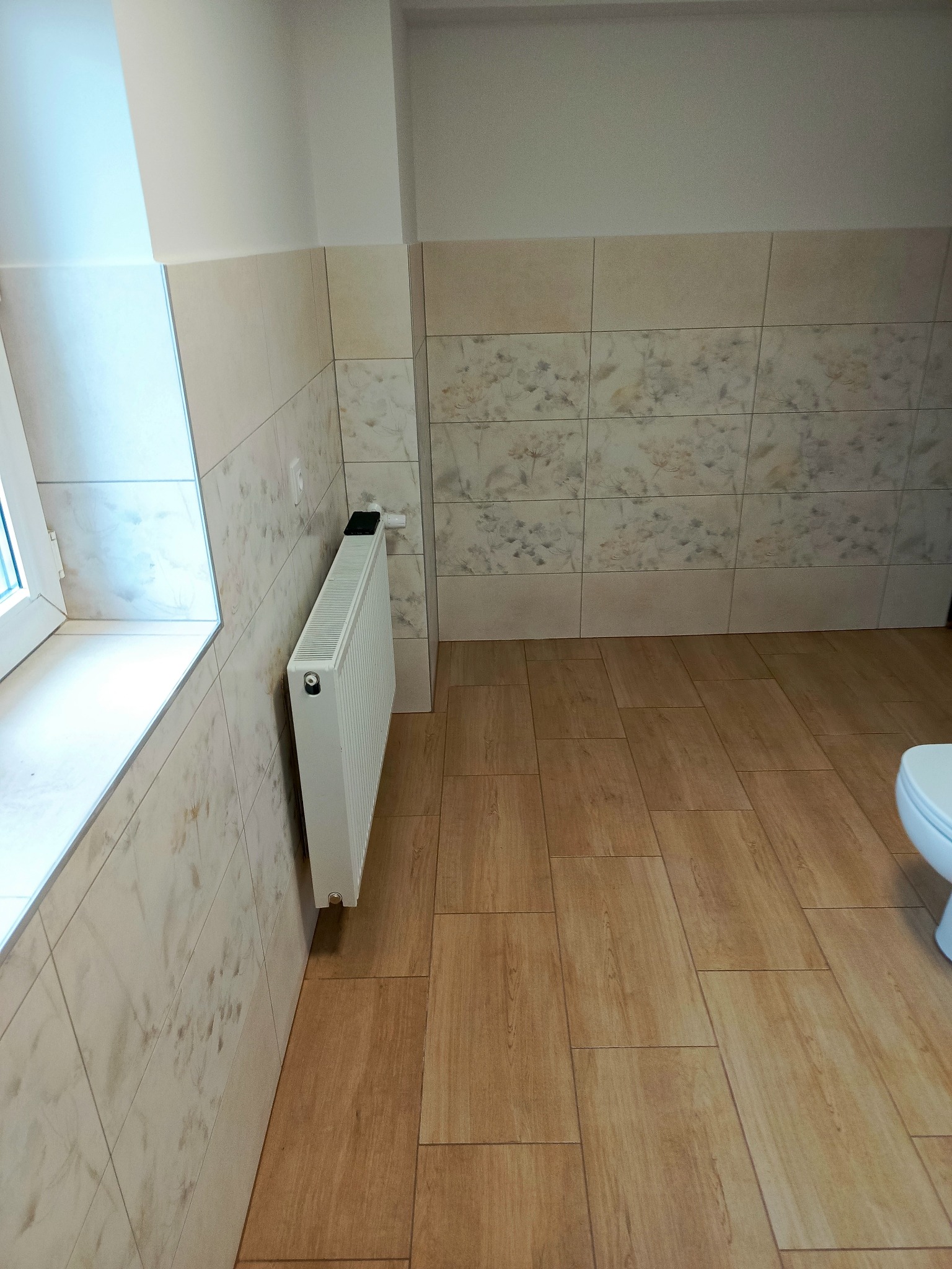 Wykończona łazienka z beżowymi płytkami imitującymi drewno na podłodze i ścianach, biały grzejnik zamontowany na ścianie, fragment toalety widoczny w rogu.