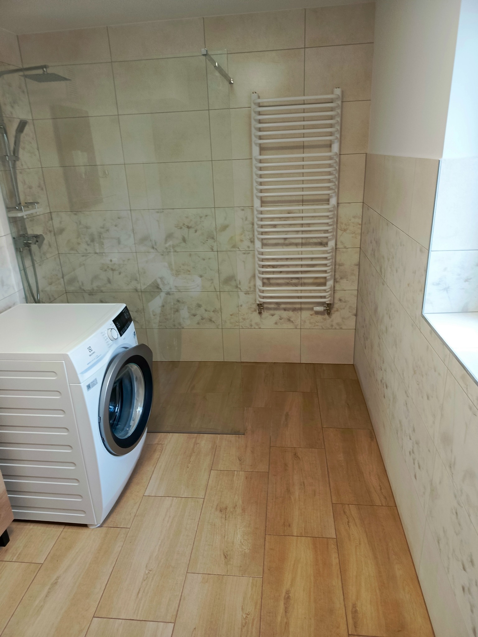 Wnętrze łazienki z kabiną prysznicową, grzejnikiem drabinkowym i pralką Electrolux, wykończone w jasnych, beżowych płytkach imitujących drewno i marmur, z widocznym odpływem liniowym w podłodze.