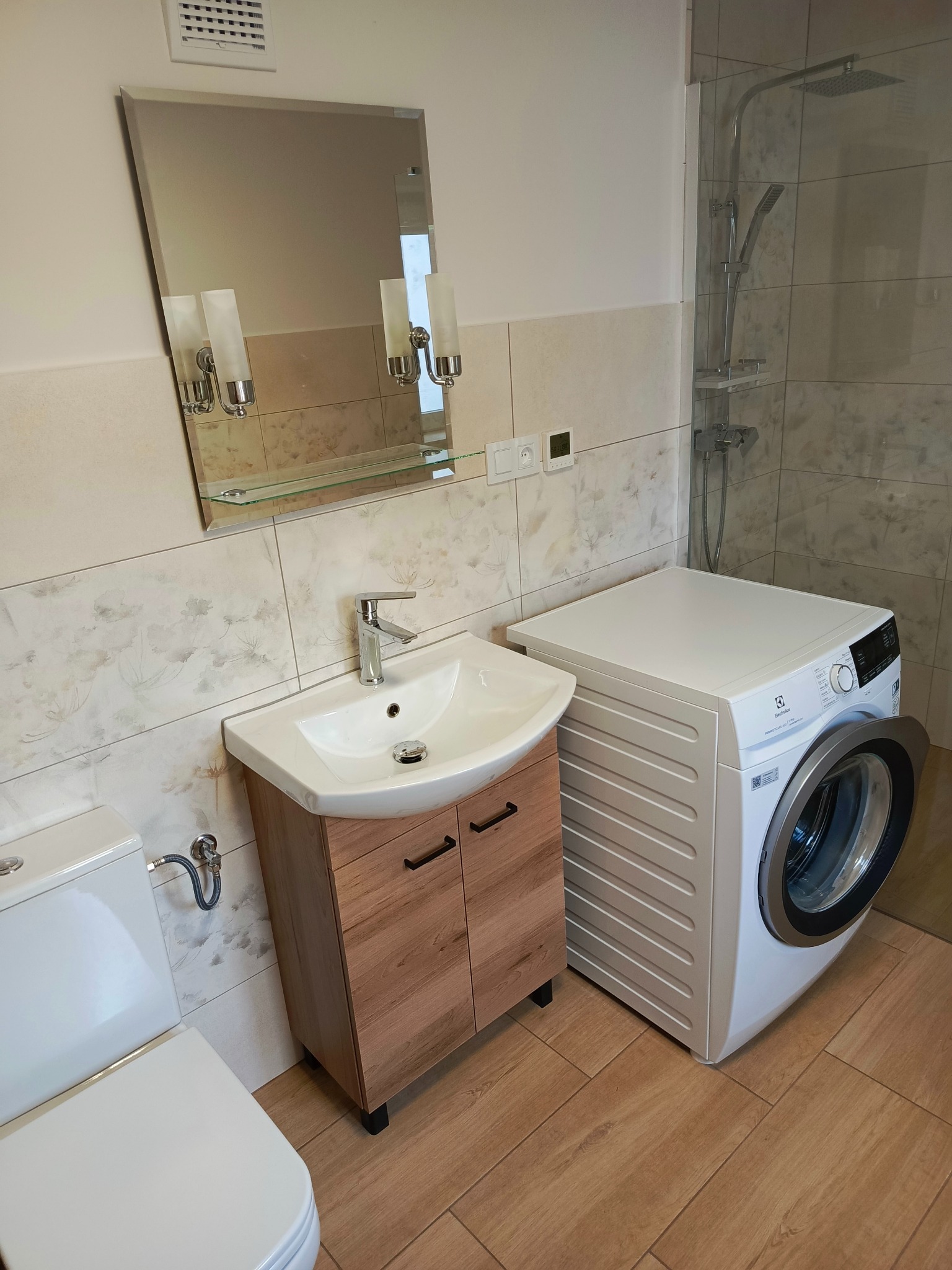 Jasna łazienka z umywalką na szafce z drewnopodobnym wykończeniem, białą toaletą, pralką Electrolux i kabiną prysznicową z deszczownicą, wykończona płytkami w delikatne kwiatowe wzory.