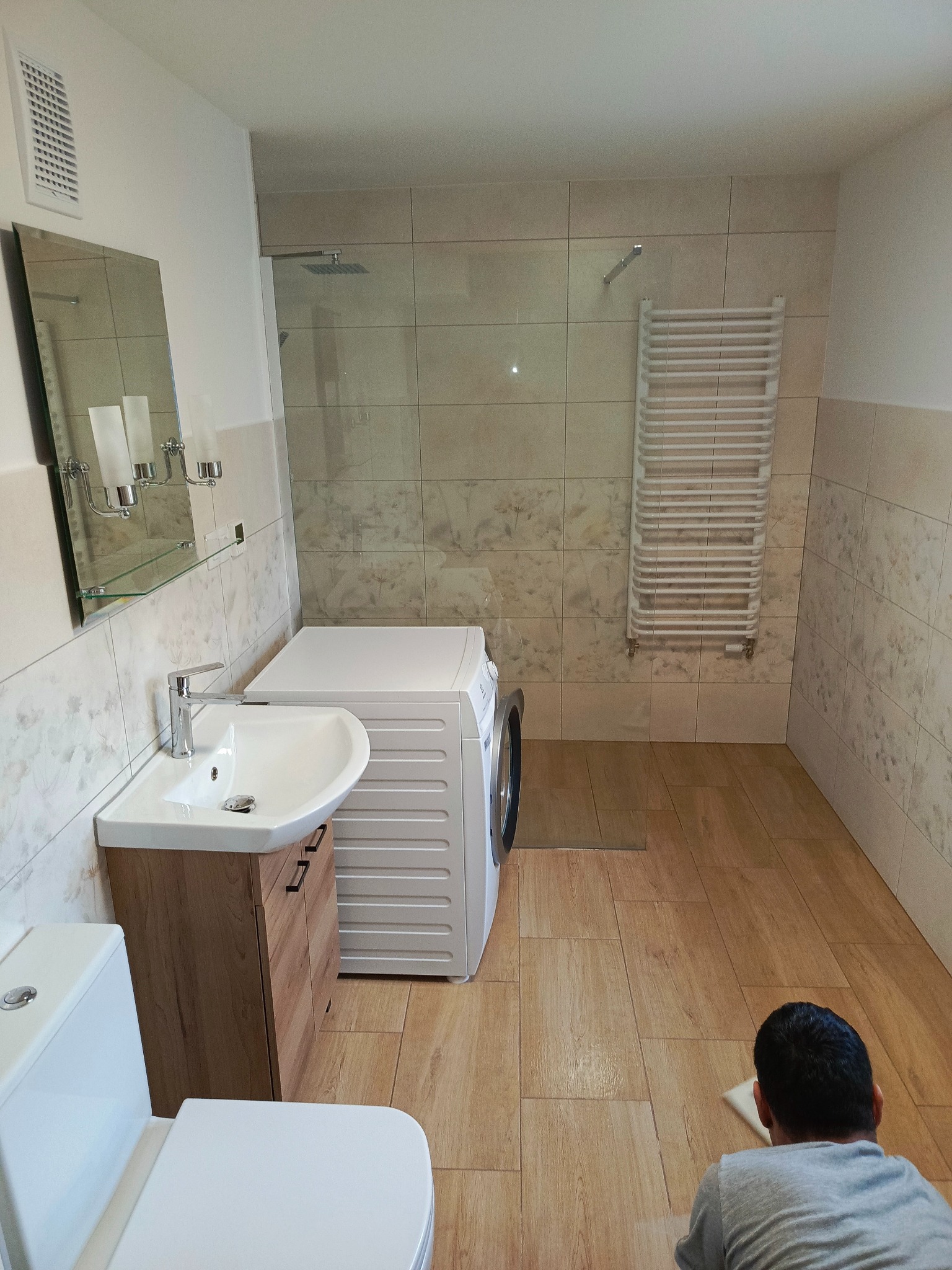 Jasna łazienka po remoncie z drewnopodobną podłogą i beżowymi płytkami, wyposażona w umywalkę z szafką, toaletę, pralkę i kabinę prysznicową z grzejnikiem drabinkowym; osoba czyści podłogę.