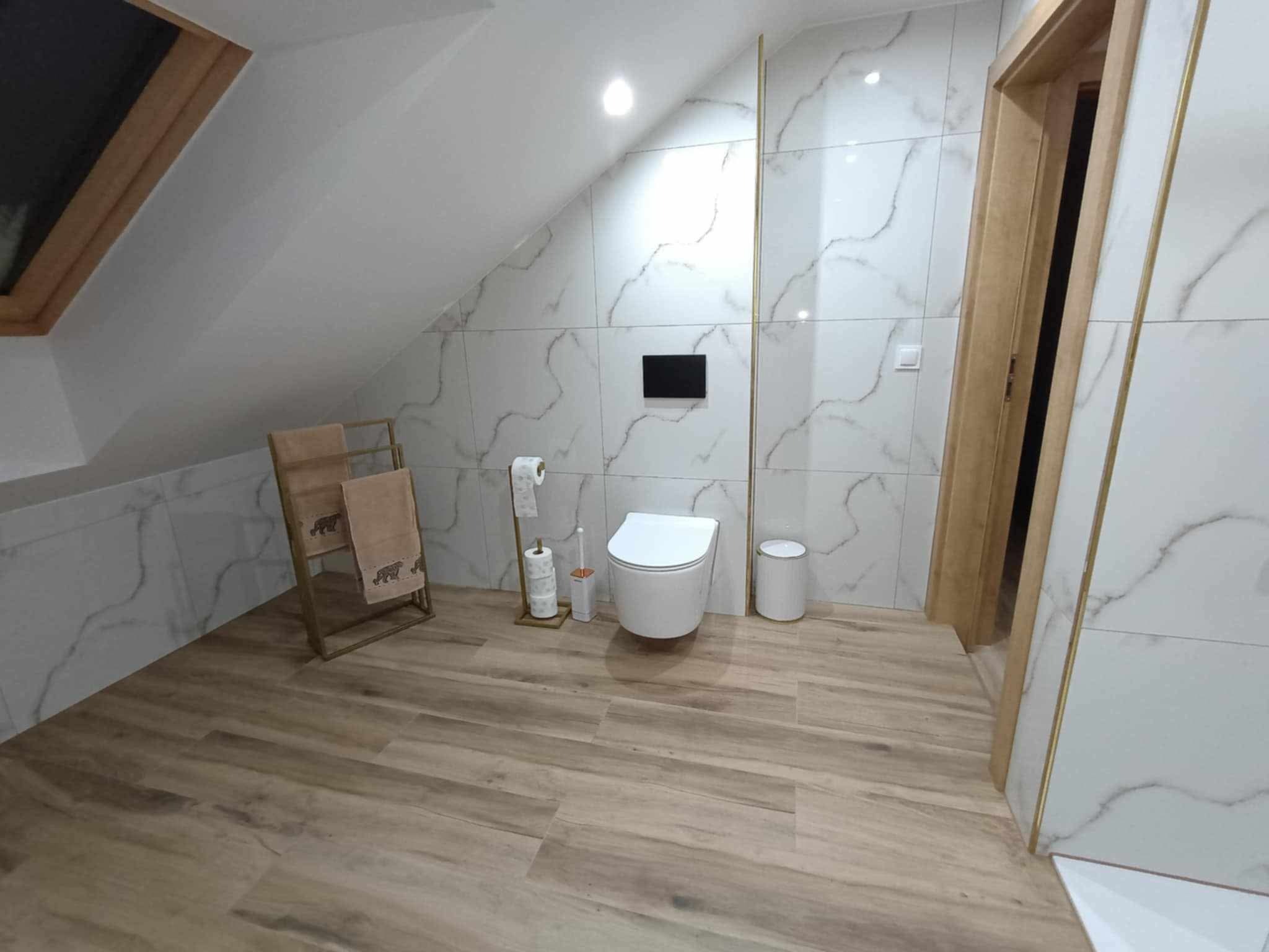Nowoczesna łazienka na poddaszu z marmurowymi płytkami, drewnopodobną podłogą i wiszącą toaletą, akcentowana złotymi listwami i dodatkami.