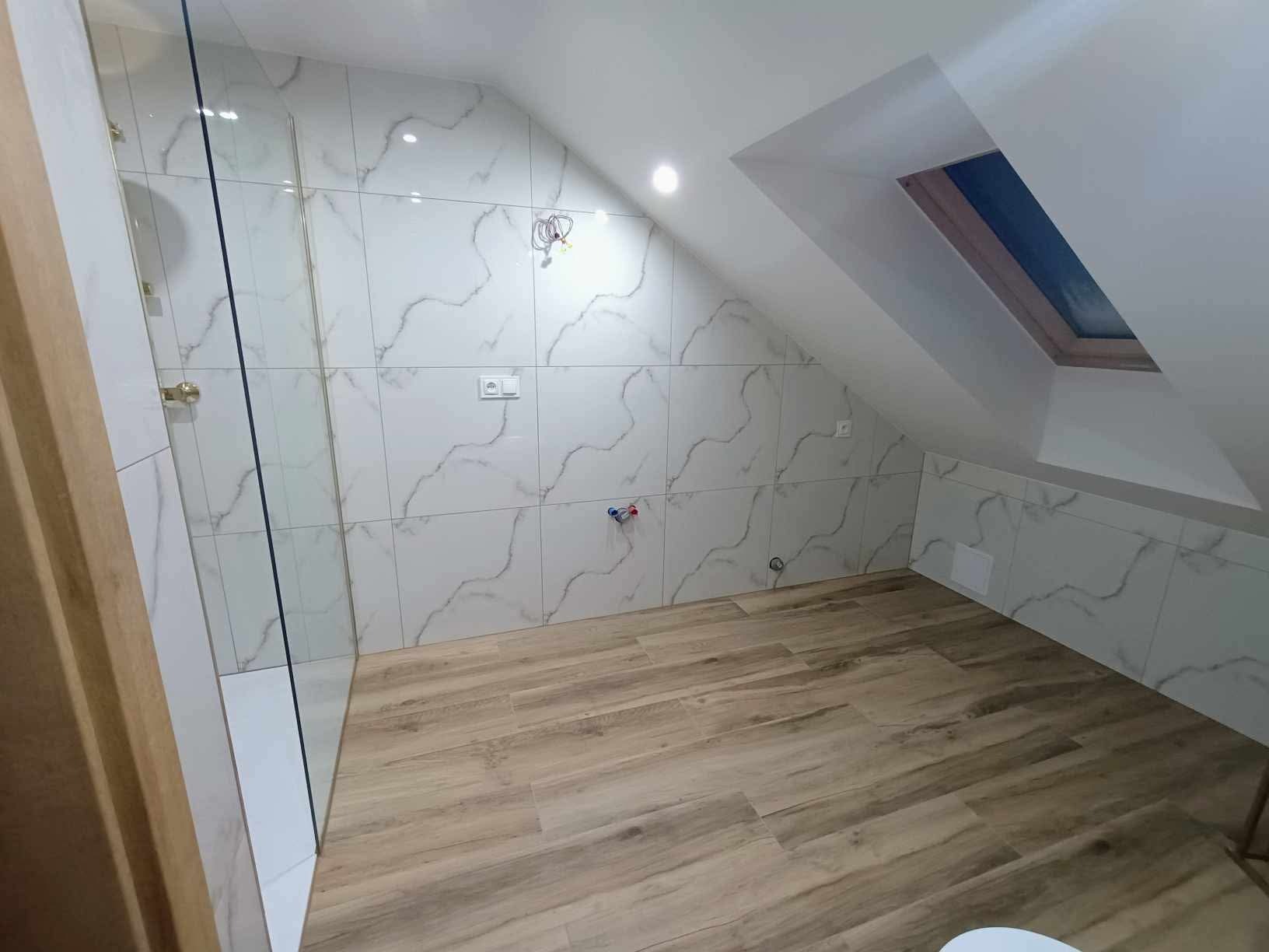 Ukończona łazienka na poddaszu z kabiną prysznicową, białe marmurkowe płytki ścienne, drewnopodobna podłoga, okno dachowe i widoczne przyłącza wodne.