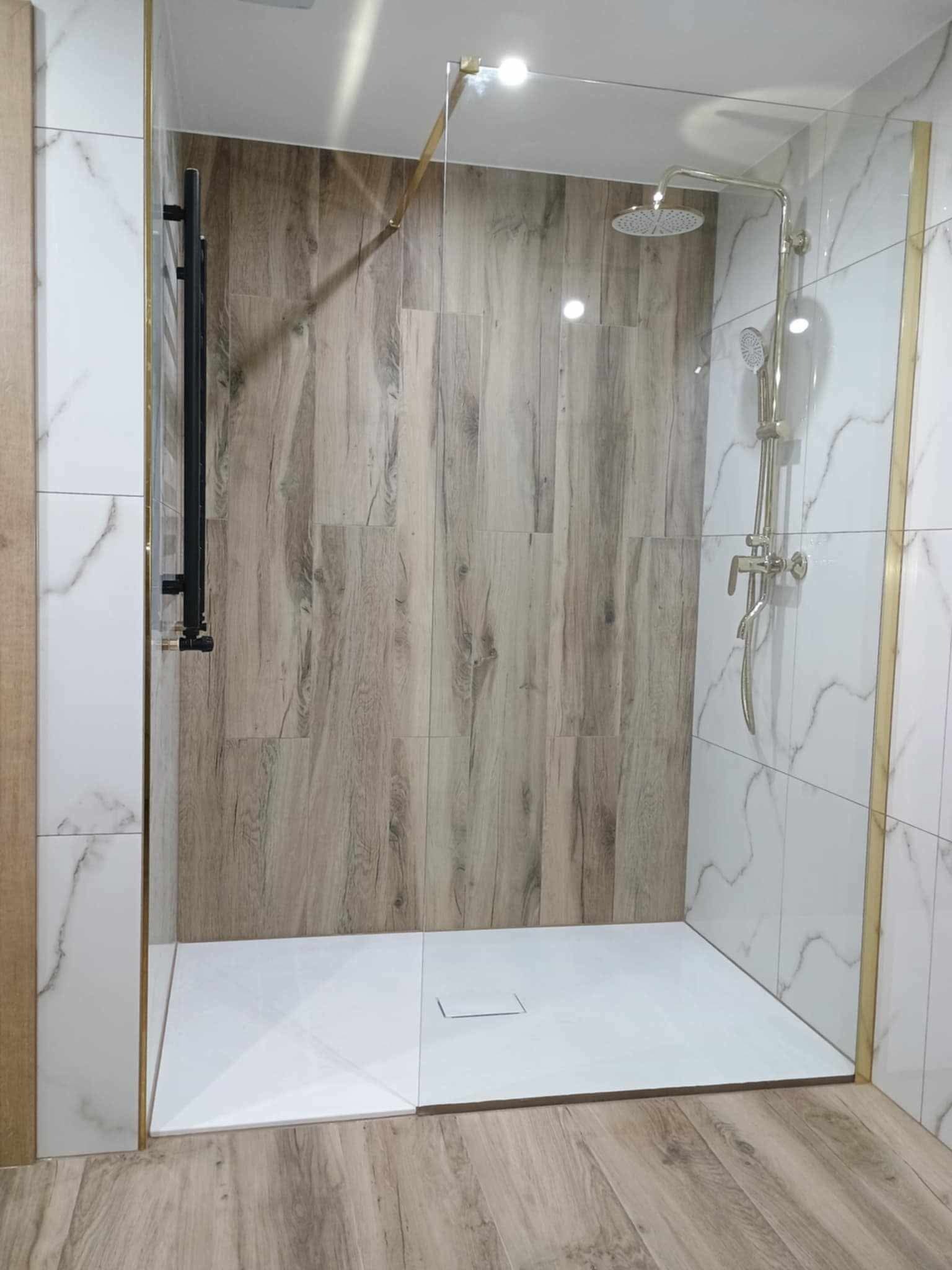 Nowoczesna kabina prysznicowa walk-in z panelami imitującymi drewno i białymi płytkami marmurkowymi, wykończona złotymi listwami, z czarnym grzejnikiem drabinkowym.